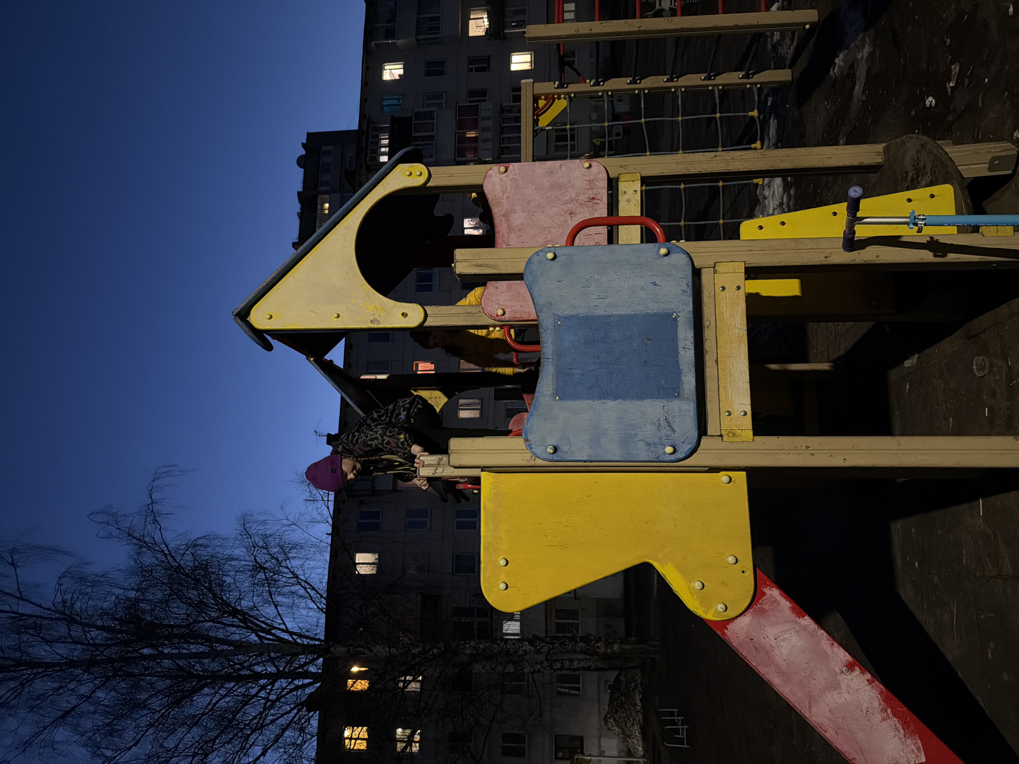 A child wearing a pink hat and patterned jacket is perched on a playground structure. The structure is made of wood and colorful plastic components in shades of yellow, blue, and red. The structure appears to be a playhouse with a slide and climbing elements. Behind the playground structure, a multi-story apartment building is visible. Some of the windows are illuminated. There are bare tree branches silhouetted against the twilight sky. The scene appears to be an evening shot. The playground is in the city of Chaikovsky, Russia.