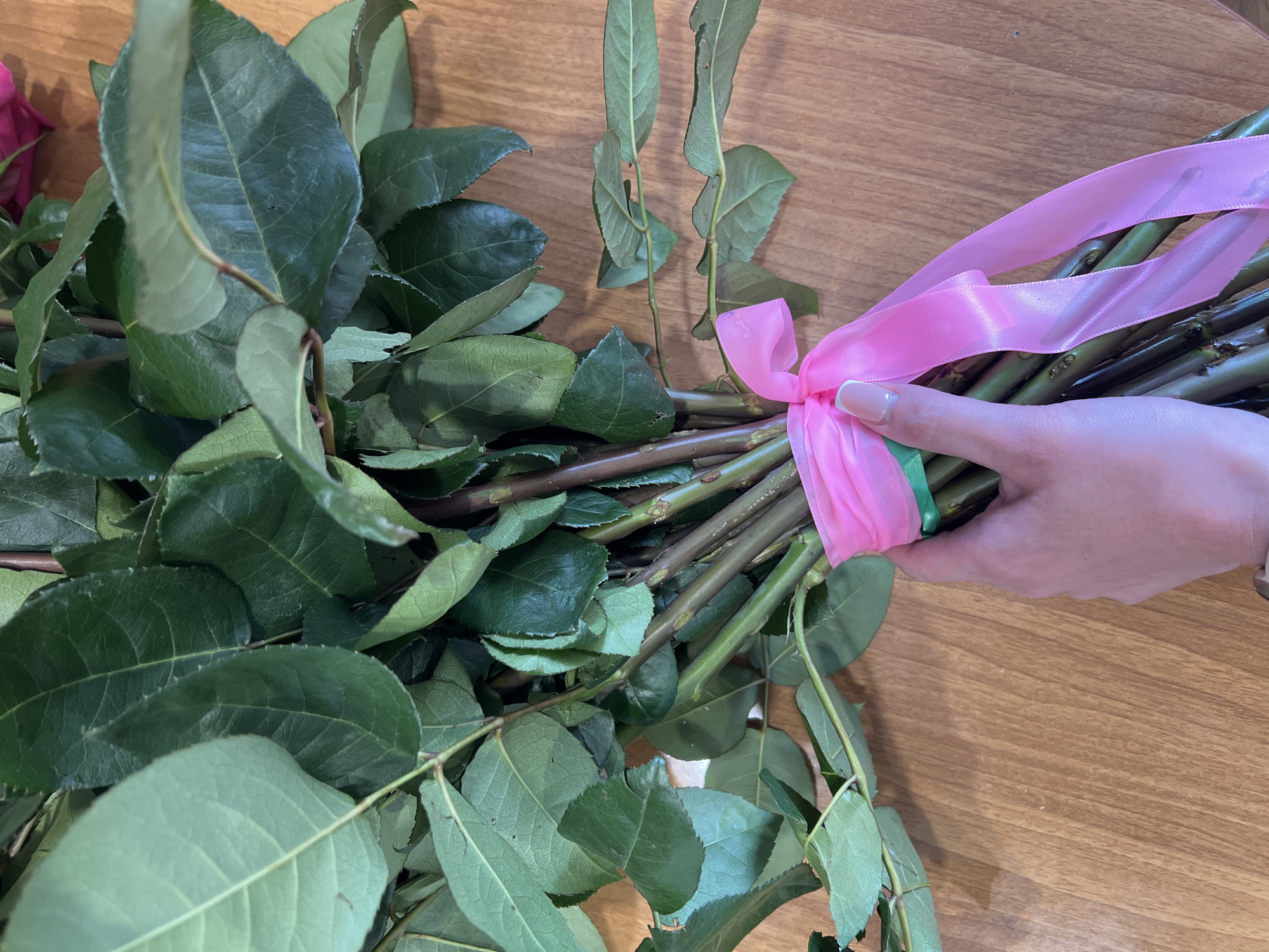 A person's hand is holding a bouquet of rose stems tied together with pink and green ribbons. The stems are surrounded by green leaves. The background is a wooden surface. The location is unknown, and there is no information to suggest this image was taken in Votkinsk, Russia.