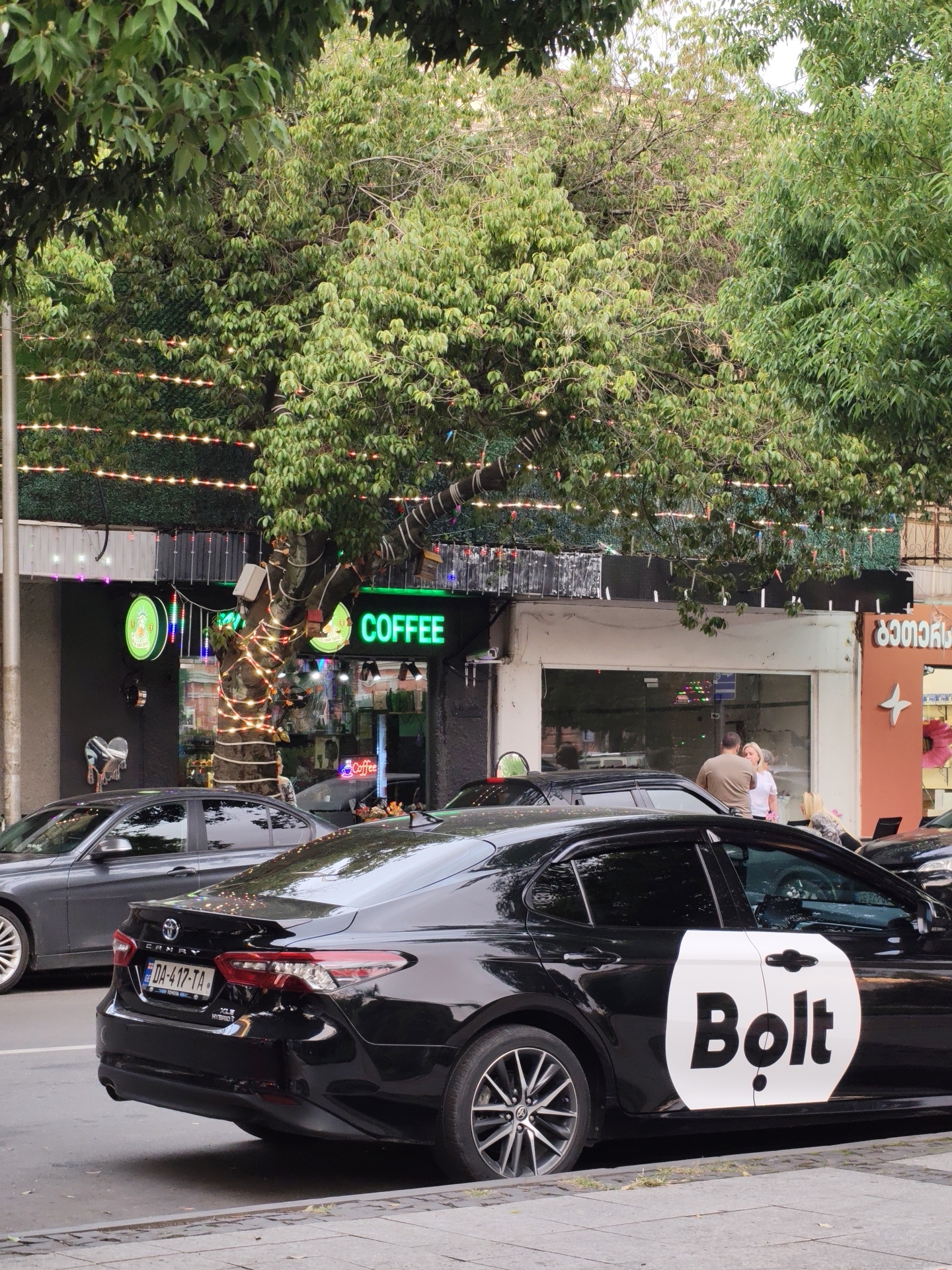 The image captures a street scene in Batumi, Georgia, with a focus on a black Bolt ride-sharing car parked at the curb. The car is a Toyota Camry Hybrid and features the distinctive white Bolt logo on its rear passenger door. In the background, a tree decorated with string lights is situated in front of a building with a "COFFEE" sign in neon green. Other vehicles, including a gray sedan, are visible on the street. A couple stands near a storefront in the middle ground, and a person with blonde hair sits on a bench to the right. The environment suggests an urban setting with commercial establishments and pedestrian areas.