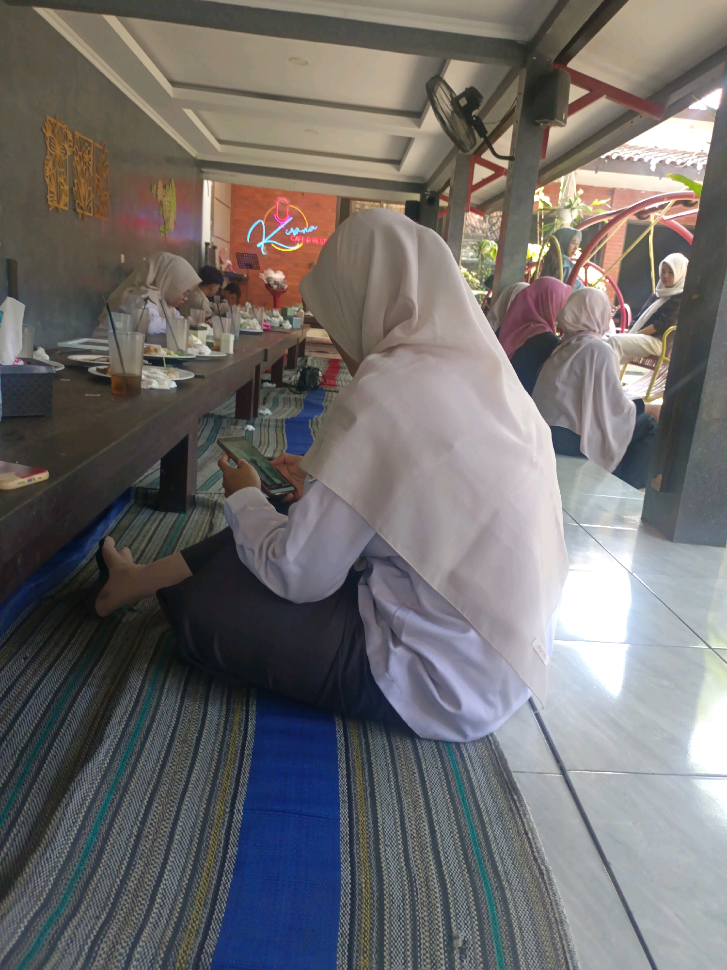 In the city of Kerangkulon, Indonesia, a cafe with an outdoor seating area is depicted. A young woman wearing a light-colored hijab and a white shirt is seated on a striped mat on the floor, looking at her smartphone. She is positioned to the left of the frame, with her legs crossed and her feet visible. In the background, other patrons are seated at a long, wooden table laden with plates of food and drinks. A neon sign with the word "Kirana" is visible on the brick wall behind them, along with a bouquet of flowers. The cafe appears to have a relaxed, informal atmosphere, with ceiling fans and red-framed support beams visible. The floor is tiled, and sunlight streams in, creating reflections.
