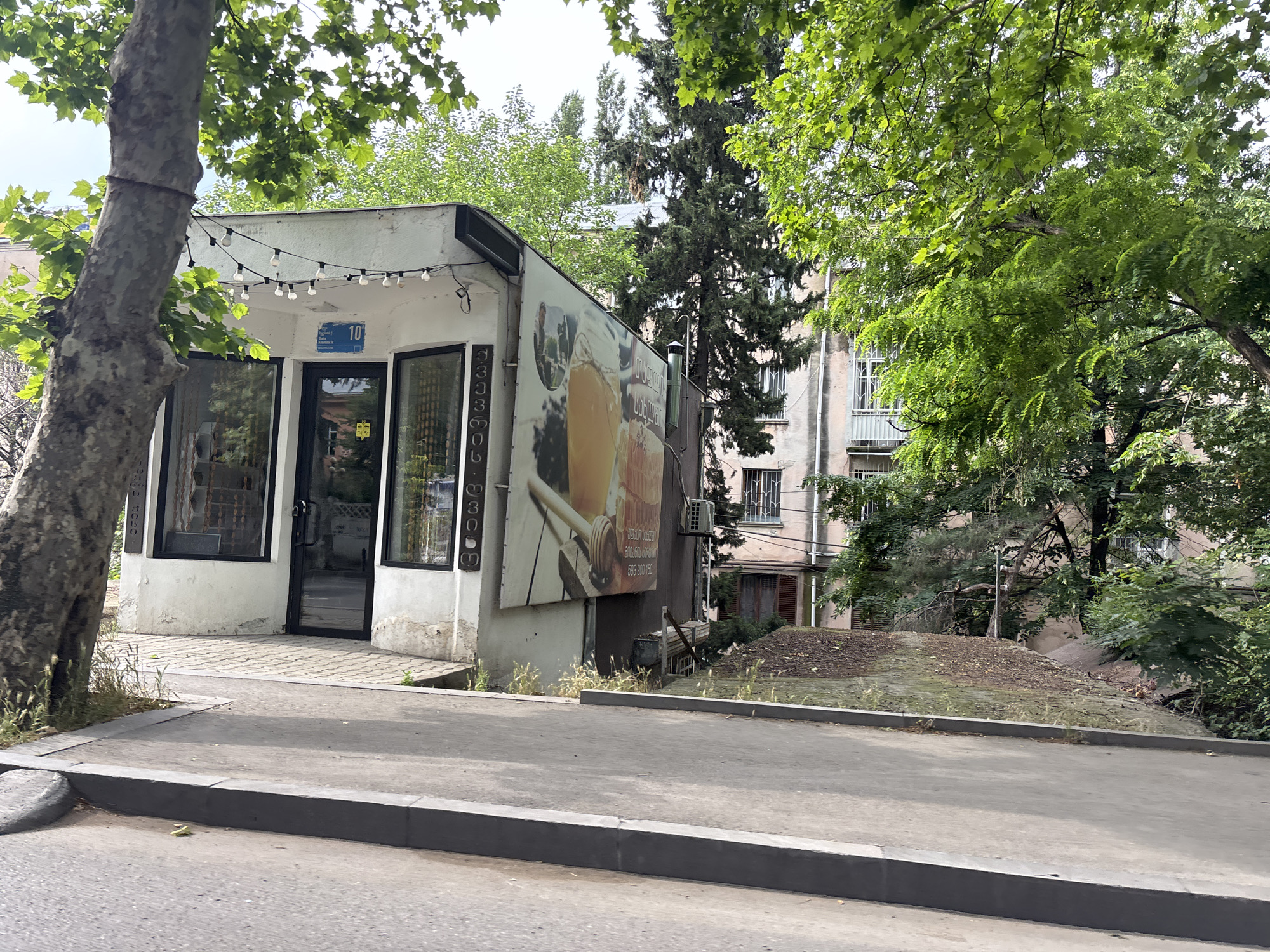 The image shows a small, white building with a flat roof, possibly a kiosk or small shop, located at number 10 on a street in Tbilisi, Georgia. The building has large glass windows and a glass door. A string of lights is hung above the entrance. A large advertisement poster is attached to the side of the building, depicting what appears to be a beverage and some text in Georgian. A large tree with green foliage is situated to the left of the building, partially obscuring it. Behind the building, a multi-story apartment building with pinkish-beige walls is visible, with several windows and balconies. The area is surrounded by greenery, with more trees and foliage present. The ground in front of the kiosk is paved with hexagonal stones, and a sidewalk runs alongside a paved street. The overall scene suggests an urban environment in Tbilisi on a cloudy day.