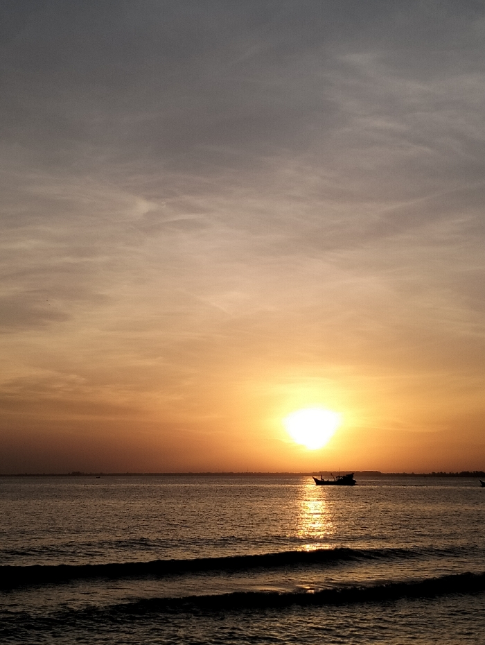 A vibrant sunset scene over the ocean. The sun, a bright orb, reflects intensely on the water's surface. A small boat silhouette is visible near the sun's reflection. The sky blends from grey to orange hues, indicative of the setting sun. Gentle waves lap the shore in the foreground. The horizon appears level with distant land visible on either side.
