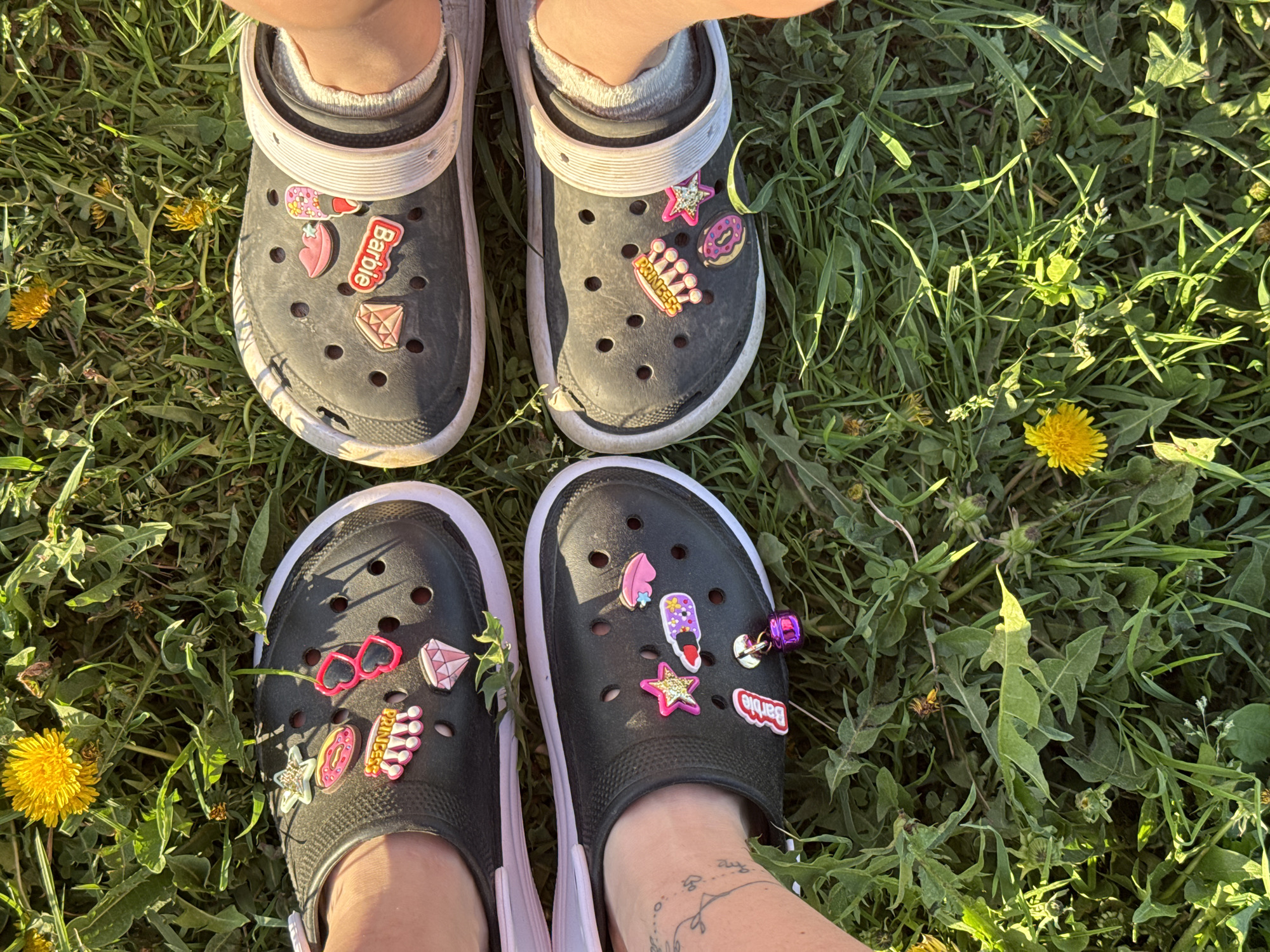 The image shows a top-down view of four feet wearing black Crocs with white soles. The Crocs are decorated with colorful charms. The feet are standing on grass with dandelions. The image does not contain information to confirm that this image was taken in the city of Rusalevka, Russia.