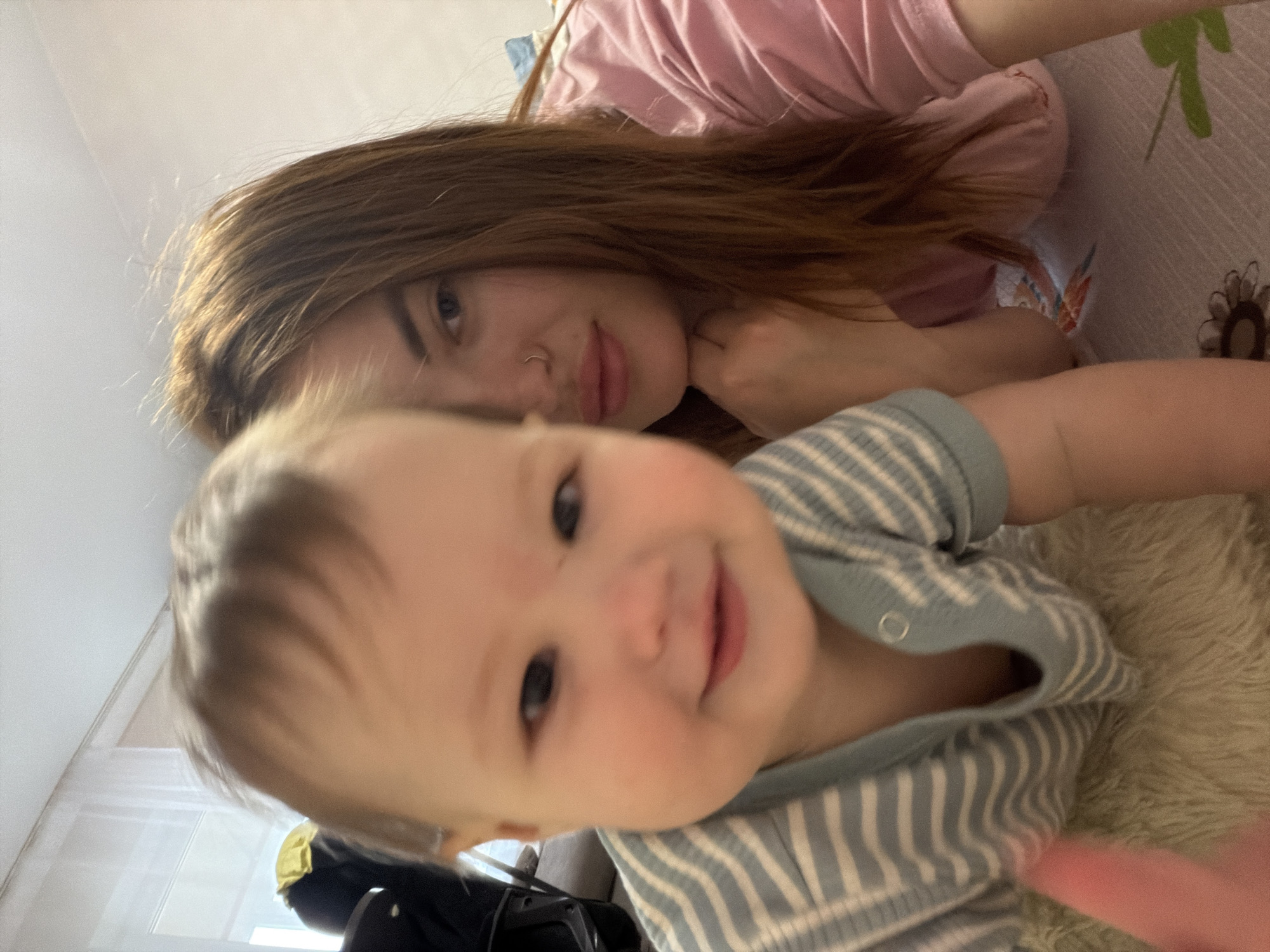 A selfie shows a young woman and a baby on a bed in Unknown, Unknown. The woman, with light brown hair and a nose ring, is wearing a pink shirt and smiling. The baby, with light skin, is wearing a striped blue and white shirt and also smiling. A furry rug is visible. There is a window in the background.