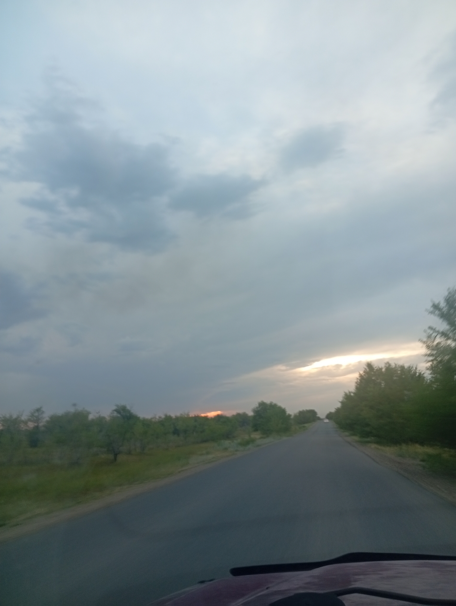 A low-angle, medium shot captures a paved road leading towards the horizon, framed by green vegetation on either side. The sky is a mixture of grey and white clouds, with a sliver of orange sunlight breaking through near the right side of the frame, suggesting it is either sunrise or sunset. The road itself is empty of vehicles, except for a distant white car barely visible in the far distance. The foreground shows the hood of a dark red vehicle, implying the image was taken from inside a car. The scene evokes a sense of travel and openness.
