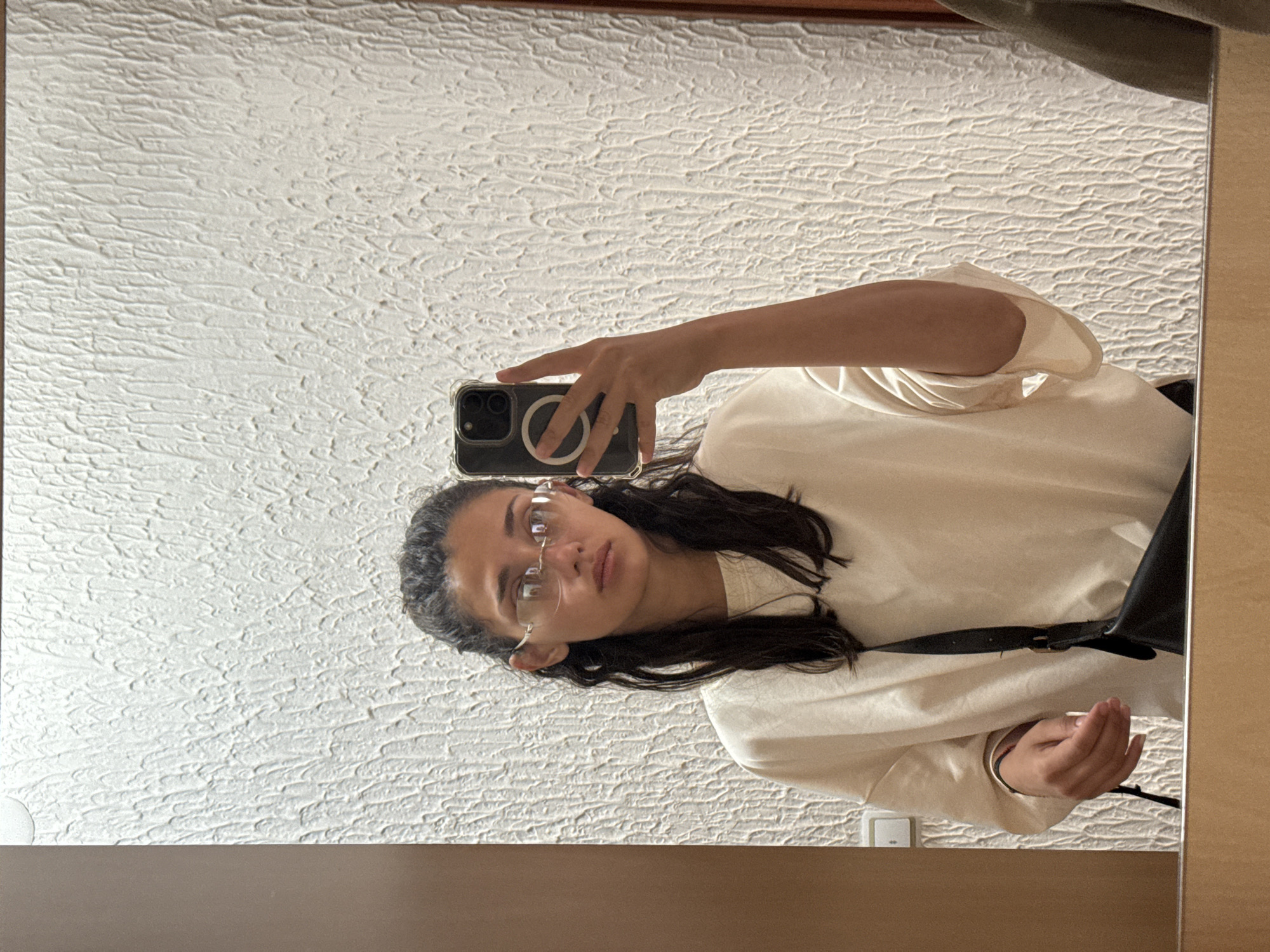 A young woman with dark, wavy hair tied back is reflected in a mirror. She is wearing a light-colored, oversized sweatshirt and rimless glasses. Her left hand is holding a smartphone, and she is taking a selfie. A black crossbody bag is visible across her body. The background of the mirror is a textured white wall. The environment appears to be indoors, possibly a bathroom or hallway, based on the wall texture and the presence of a switch on the wall. The location is identified as Miljevci, Montenegro.
