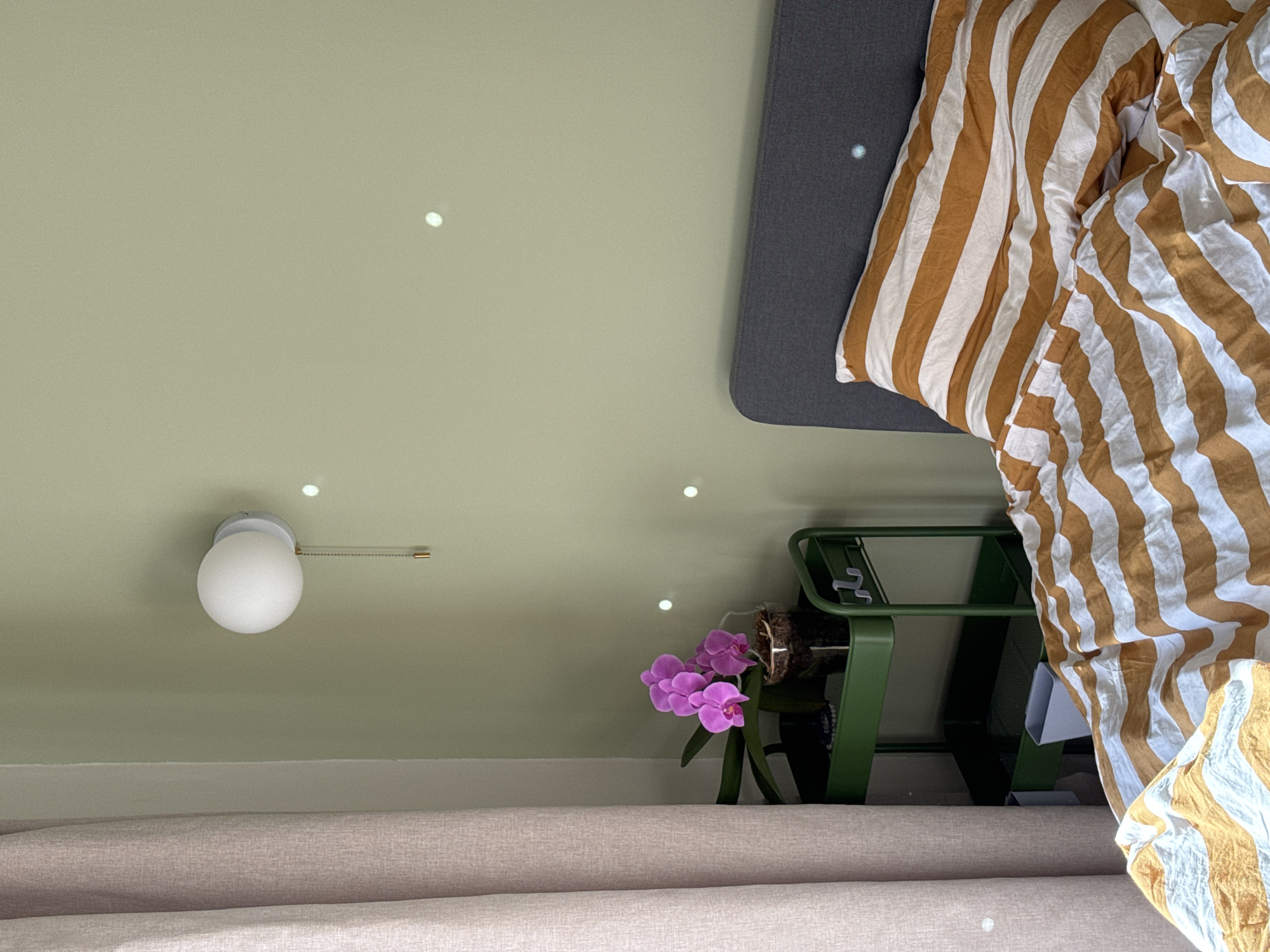 The image depicts a bedroom scene with a bed, a nightstand, a curtain, and a wall lamp. The bed is made with a comforter and a pillow, both featuring a striped pattern of orange and white. To the left of the bed, there is a dark gray headboard and a green metal rolling cart acting as a nightstand. On the top shelf of the cart, a potted orchid with pink flowers is visible. A pale pink curtain hangs on the left side of the image. On the light green wall, a white globe-shaped wall lamp with a pull chain is mounted, and several small, circular white specks are scattered across the wall, possibly reflections or dust particles. The overall impression is that of a softly lit, contemporary bedroom. The location is identified as Izhevsk, Russia, although no specific visual cues within the image directly confirm this.