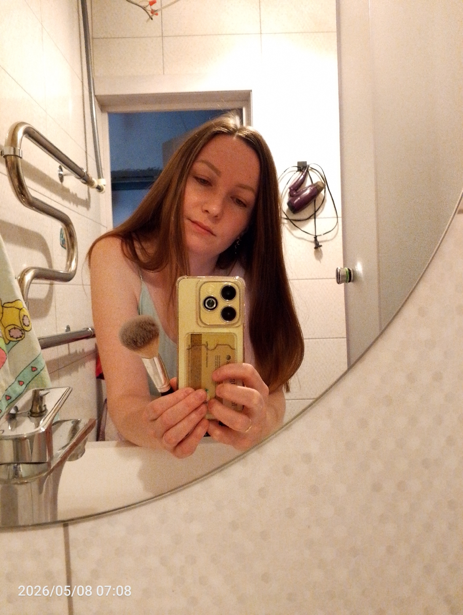 A woman is taking a selfie in a bathroom. The reflection is in a round mirror positioned above a white sink with a silver faucet. The woman is holding a makeup brush in one hand and a yellow smartphone in the other, obscuring part of her face. She has long brown hair and is wearing a light-colored top. The bathroom features a tiled wall, a towel rack, and a hair dryer mounted on the wall with cords. The photo includes a timestamp of "2026/05/08 07:08." There is insufficient information to determine the specific location in the city of Chaikovsky, Russia.