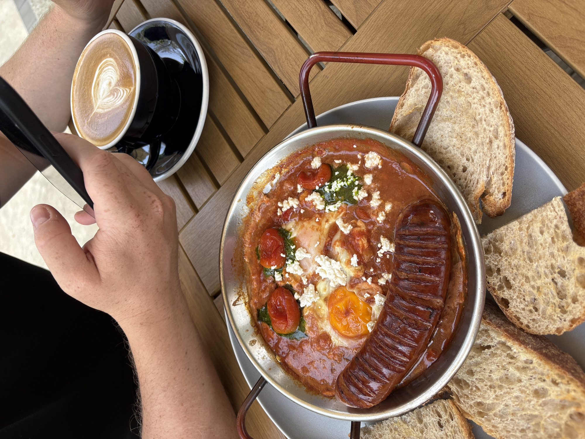 A top-down shot depicts a person seated at a wooden table, holding a smartphone in their left hand. A black coffee cup sits on a black saucer to the left of the frame. In the center, a metal pan containing shakshuka, sausage, feta cheese, tomato and pesto is presented on a grey plate alongside slices of bread. The location is inferred to be Choeng Thale, Thailand. The individual seems to be interacting with the smartphone.