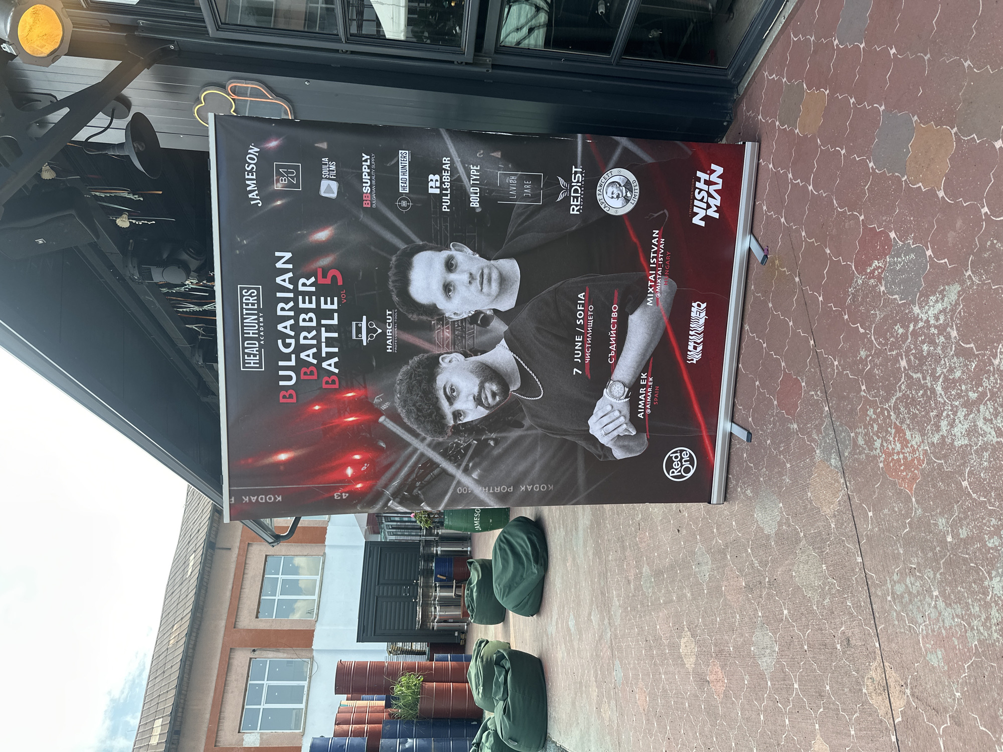 An outdoor scene in Sofia, Bulgaria, features a large vertical banner promoting the "Bulgarian Barber Battle 5 VOL" event. The banner, positioned on a stand, displays two male barbers: Aimar Ek from Spain and Mixtai Istvan from Hungary. Event details on the banner indicate the date "7 JUNE" and location "SOFIA / ЧИСТИЛИЩЕТО" (Purgatory). Numerous sponsor logos are visible, including Jameson, Pull&Bear, Red One, and Nish Man. To the left of the banner, a collection of stacked red and blue industrial barrels, some repurposed as planters, lines the perimeter of the area. Several dark green beanbag chairs are scattered across the reddish-brown paved ground, which features a repeating quatrefoil pattern. In the background, a light-colored building with a tiled roof and multiple white-framed windows is visible. To the right, an industrial-style overhead structure equipped with a speaker, a dark hanging light, and an orange spotlight is attached to a building wall. The sky, partially cloudy, is visible in the upper left corner.