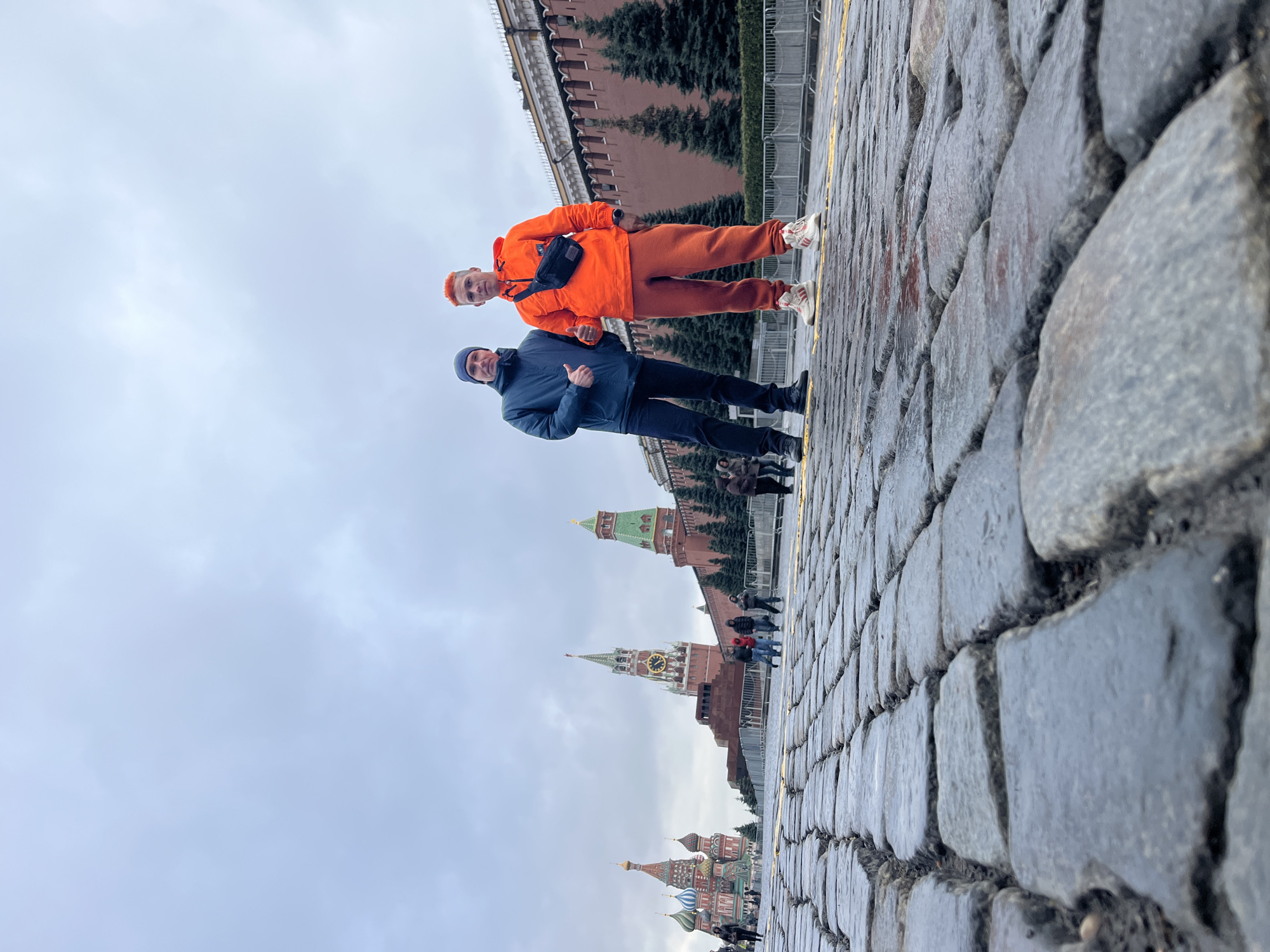 Two people are standing on a cobblestone surface. One person is wearing a bright orange jacket and pants with white sneakers. The second person is wearing a navy blue jacket and pants. In the background, there are the walls and towers of the Moscow Kremlin.