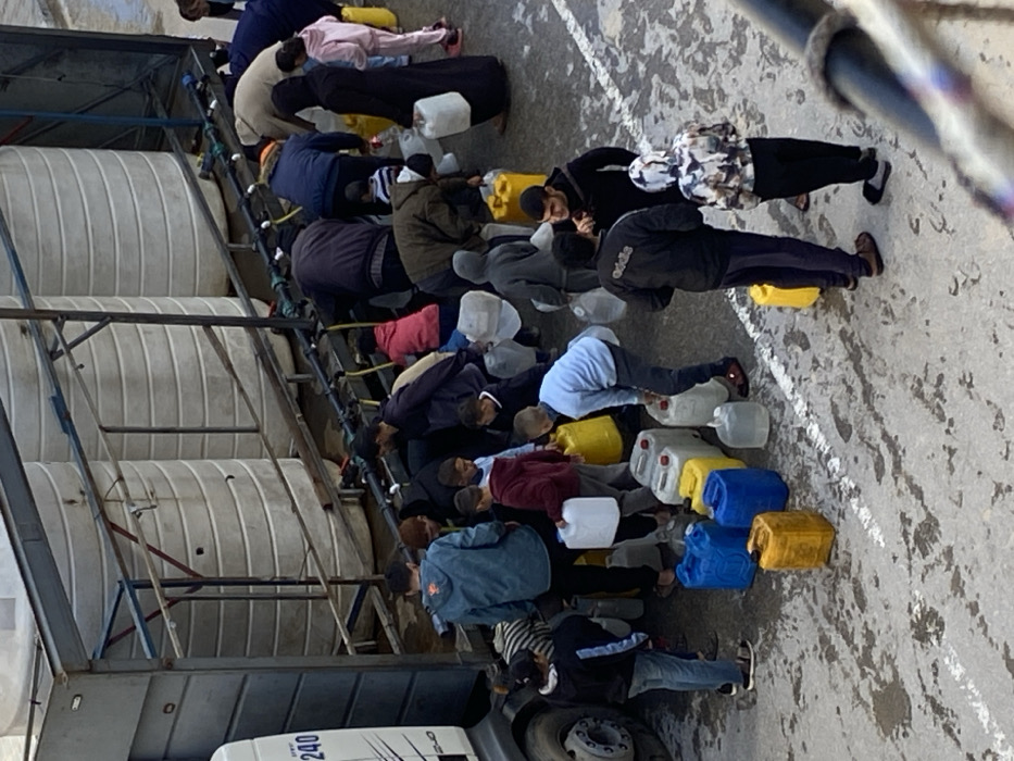 An overhead view captures approximately 25-30 individuals, comprising adults and children of varying ages, clustered around a water distribution truck in Khan Yunis, Palestinian Territory. The truck's trailer features a metal framework supporting at least four large, white, vertically oriented cylindrical water tanks. Multiple black hoses fitted with spigots extend from the base of these tanks. Numerous people are actively bent over, operating the spigots to fill an assortment of plastic containers, including clear, white, blue, and yellow jerrycans and bottles. Other individuals are observed waiting with empty containers or carrying filled ones away from the distribution points. The ground is a damp, light grey paved surface, marked by irregular darker stains and a distinct white painted line running horizontally across the mid-ground. The cab of the truck is partially visible on the left, displaying "240" on its side.