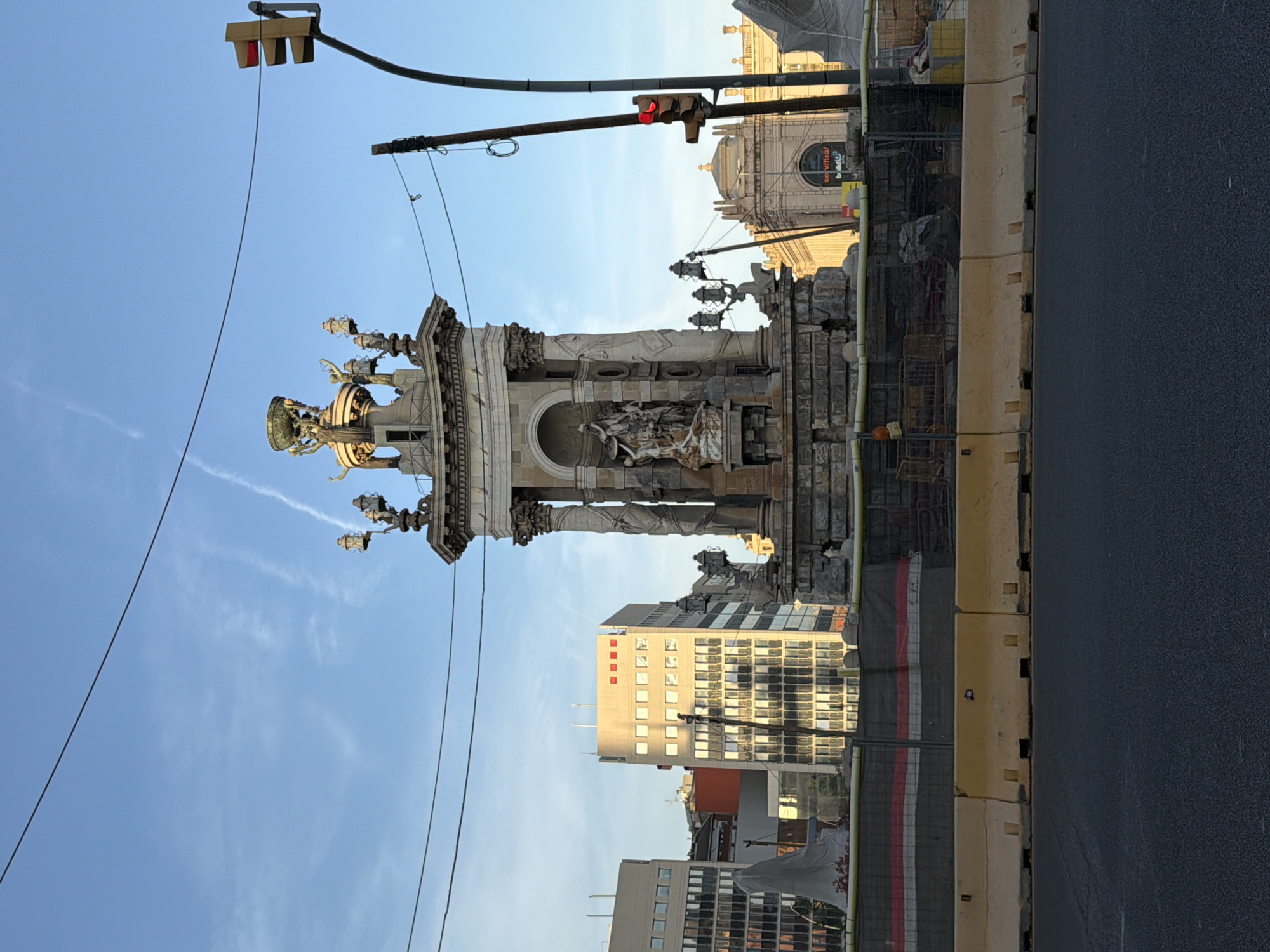 The image captures a view of the Fountain of Diana at the Palau Nacional in Barcelona, Spain. The fountain is a prominent stone structure with sculptures, and several lamps are visible. In the background, modern buildings stand, including one with a façade of windows and another with a reddish hue. A traffic light with a red signal is positioned on the right side of the frame, and overhead wires crisscross the sky. Construction barriers, made of concrete and fencing, are in place in the foreground, suggesting ongoing work or renovation. The sky is blue with faint contrails. The overall scene depicts an urban environment with a mix of historical architecture and contemporary infrastructure.