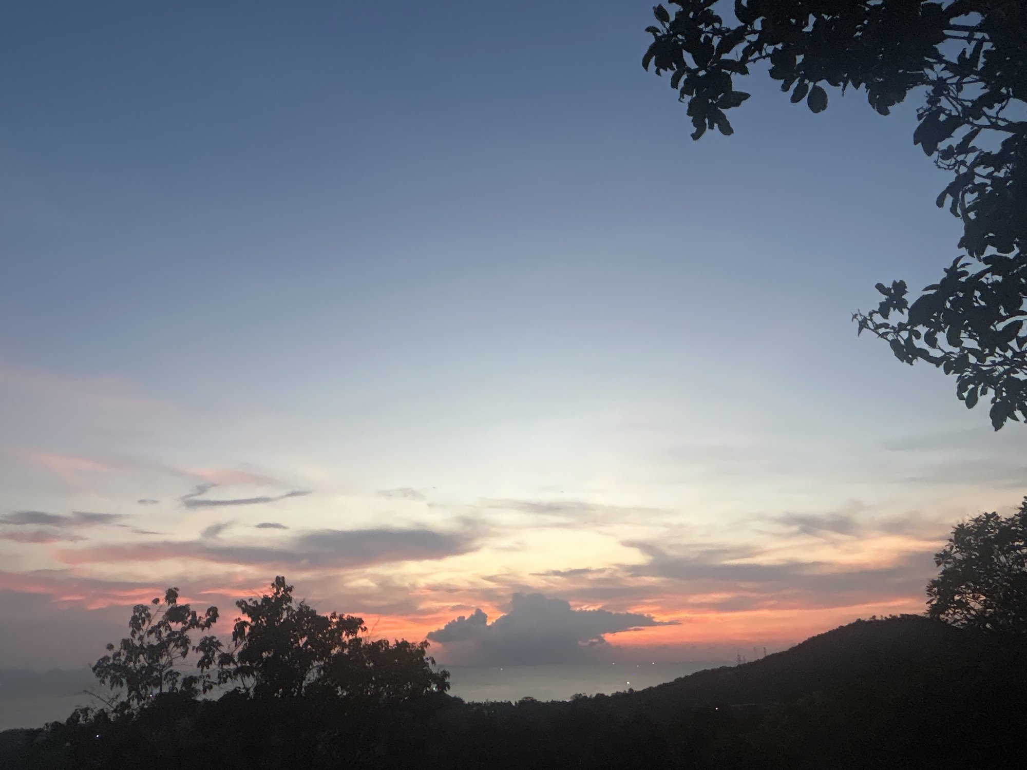 The image depicts a sunset in Baan Hin Kong, Thailand. The sky transitions from blue at the top to pink and orange hues near the horizon. There are clouds, with a prominent gray one centrally positioned. Silhouetted trees and a hill line the bottom of the image. Tree branches and leaves are visible in the top right corner.