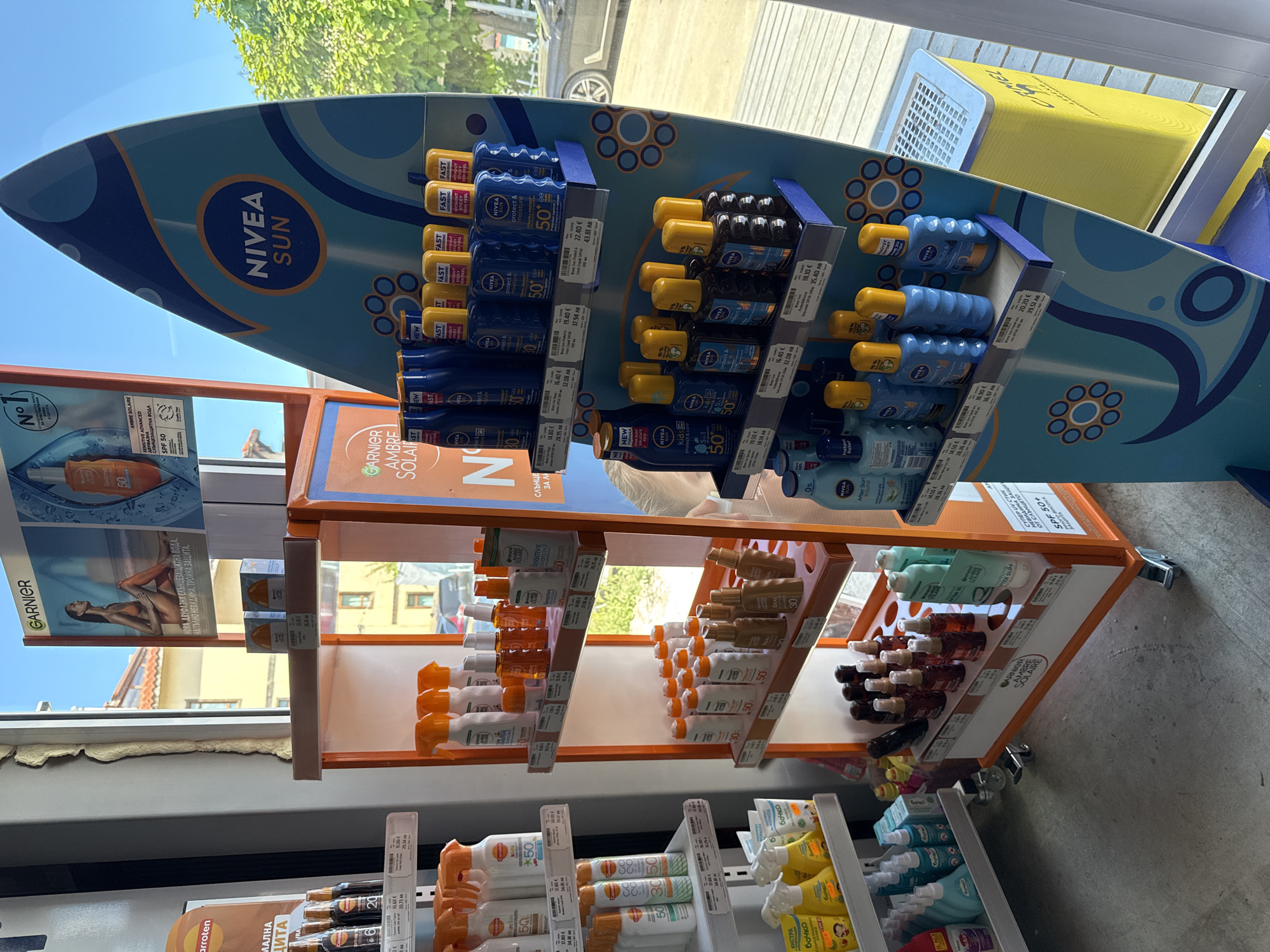 The image depicts a retail display featuring a variety of sunscreen products. The display is located in a store, likely a pharmacy or a supermarket, in the city of Tsarevo, Bulgaria. The primary focus is on a surfboard-shaped stand showcasing Nivea Sun products, including various bottles and sprays with SPF ratings. To the left, there are additional shelves with Garnier and other brands of sun protection. The products are neatly arranged on orange and blue shelves, with some products still in their packaging. The lighting suggests it is daytime, with natural light entering from an open doorway or window in the background, revealing a glimpse of buildings and trees outside. The flooring appears to be concrete. No people are clearly visible in the immediate vicinity of the display, though a person's arm and shoulder can be seen in the background, suggesting shoppers might be browsing further within the store.