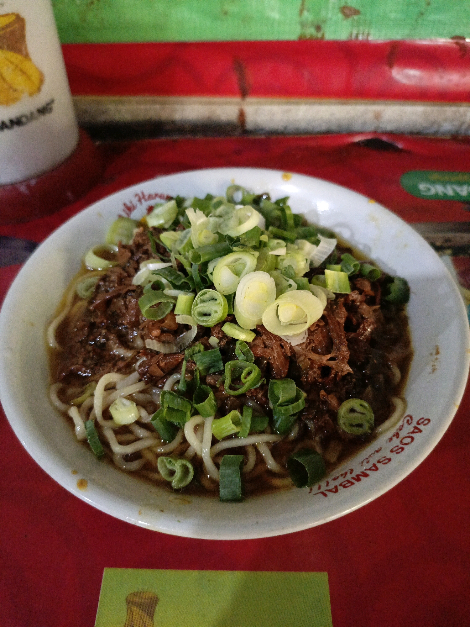 A close-up, overhead shot captures a bowl of savory noodle soup, likely Mie Ayam, a popular dish in Indonesia. The noodles are submerged in a rich, dark broth, topped with shredded chicken or meat, and generously garnished with chopped green onions and what appear to be thinly sliced shallots. The white ceramic bowl has red text around its rim, partially obscured by the noodles and toppings. The bowl rests on a vibrant red surface, possibly a tablecloth. In the background, a weathered, multi-colored surface, featuring horizontal stripes of green and red, suggests an outdoor or casual dining setting. To the left of the bowl, a portion of a white container with an illustration of a mango and text that reads "MANDANG" is visible. The overall scene evokes a sense of a humble, local eatery in Sraten, Indonesia, where this dish is commonly served.
