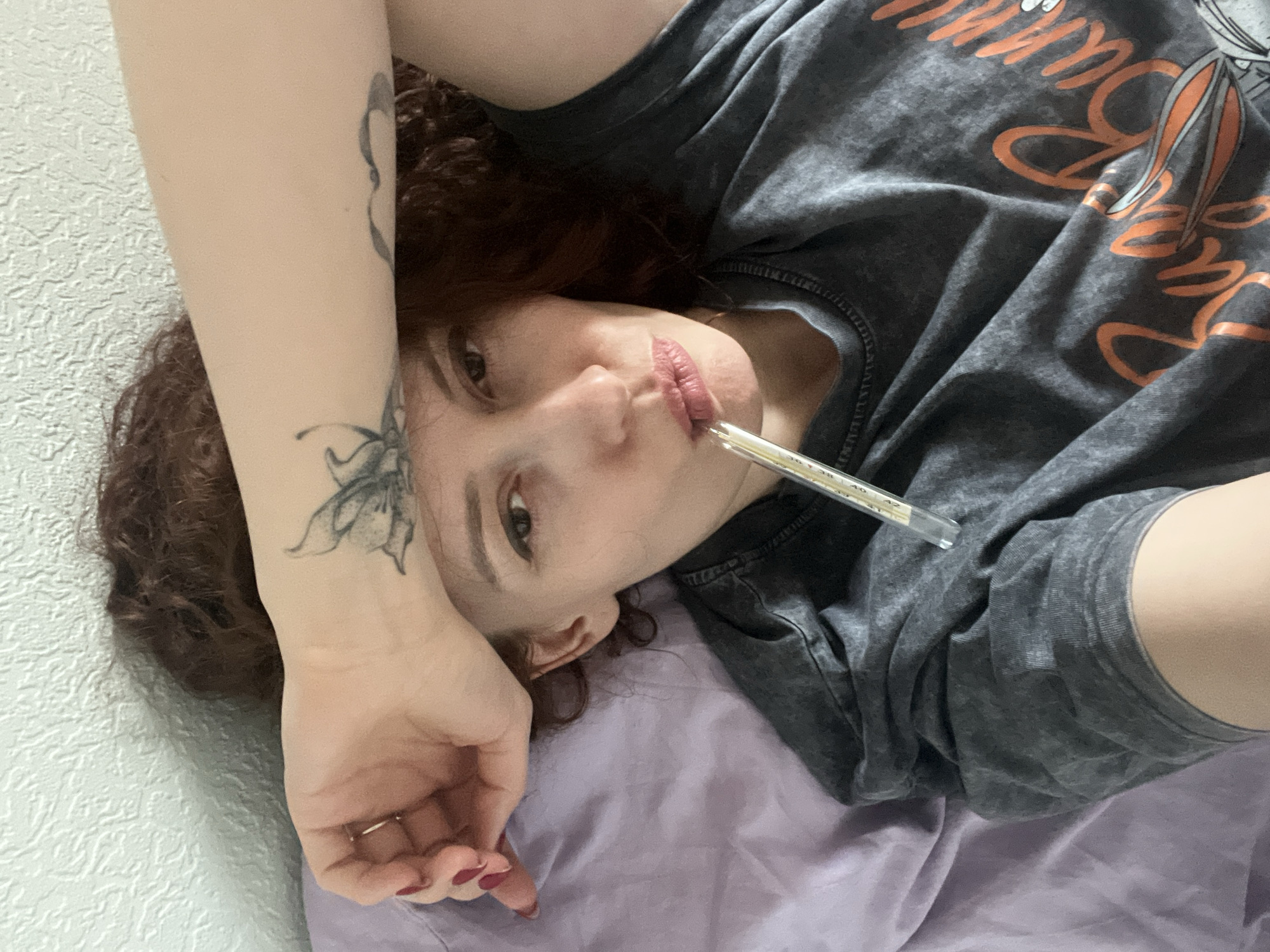 A medium close-up, eye-level shot captures a person lying down, looking directly at the camera in what appears to be an indoor setting in Saint Petersburg, Russia. The person, with reddish-brown curly hair, is wearing a dark grey, washed-out t-shirt featuring a partially visible orange graphic of "Bugs Bunny." A mercury thermometer is held in their mouth, with the mercury level indicating a temperature around 38-39 degrees Celsius, suggesting a fever.
The person's left arm, adorned with a black outline tattoo of an insect (possibly a butterfly or moth) on the forearm and another partial tattoo higher up, rests on a textured white wall. Their right arm is mostly covered by the t-shirt. The person's nails are painted a dark red or burgundy, and a simple gold ring is visible on one finger of their left hand. They are lying on light purple/lavender bedding, possibly a sheet or blanket. The overall expression is neutral, perhaps hinting at discomfort or weariness due to illness. The lighting is soft and even, typical of an indoor environment.