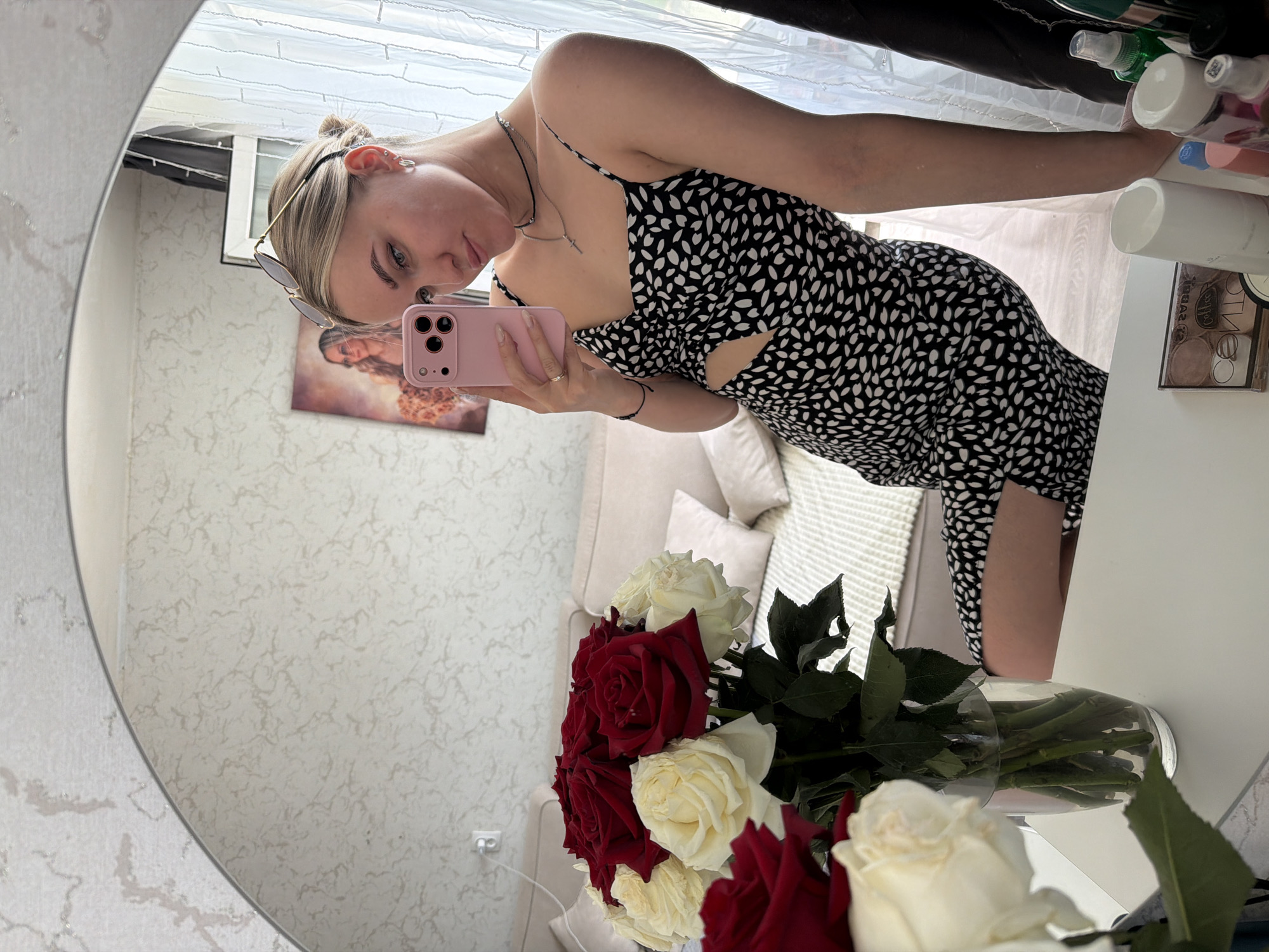 A woman with fair skin and blonde hair tied back in a bun is reflected in a large, round mirror. She is holding a pink smartphone up to her face, likely taking a selfie. She is wearing a black and white patterned dress with a cutout at the waist, along with a necklace and a bracelet. Sunglasses are placed on top of her head.
Behind her, a glimpse of an interior setting is visible, including a sofa with pillows and a piece of art on the wall. In front of the mirror, a vase filled with a bouquet of red and white roses is placed on a table or surface. Various cosmetics and personal care items are visible in the upper right corner of the frame. The background includes a window covered by blinds.