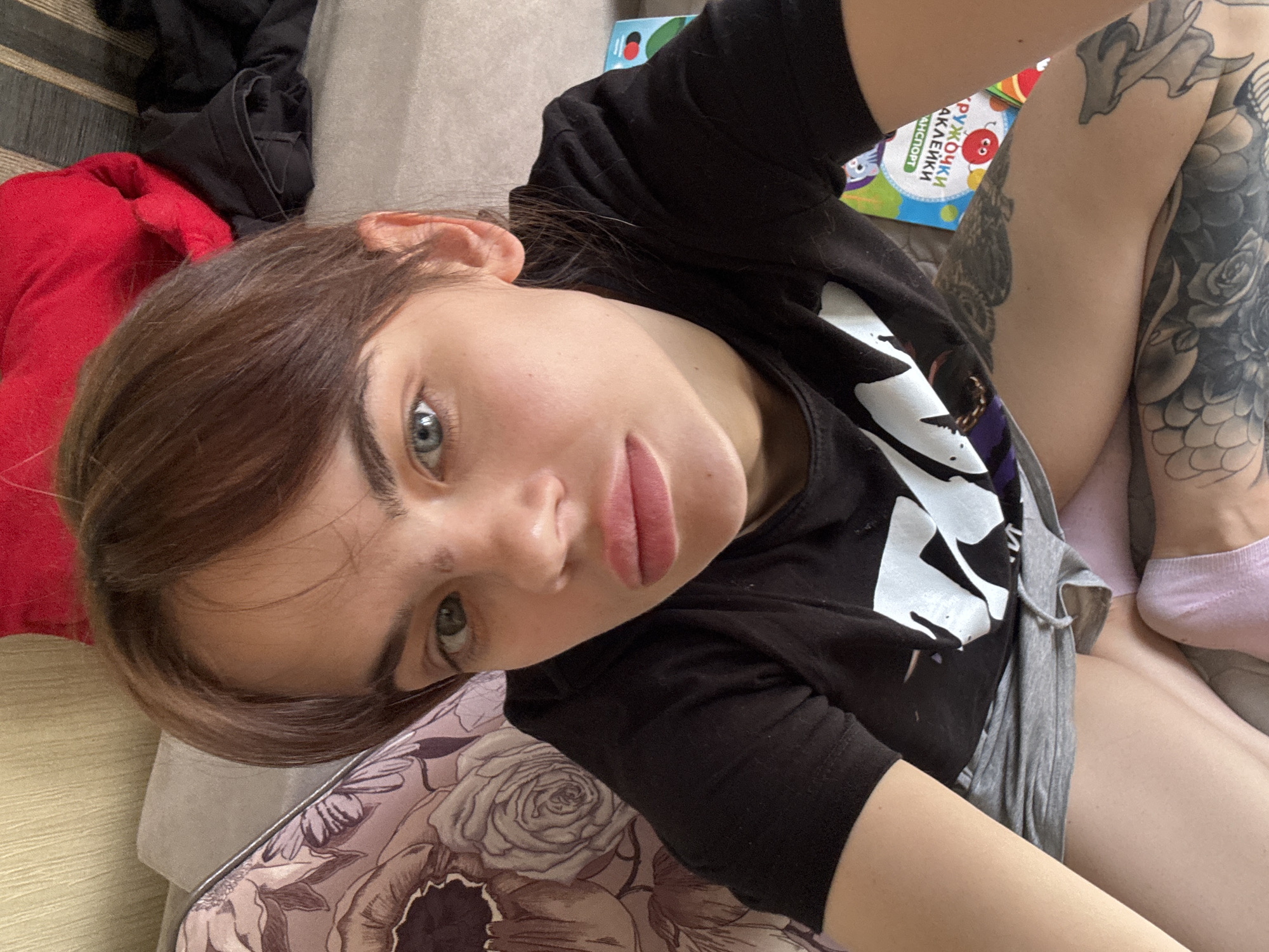 A young woman is taking a selfie while lying on a patterned cushion in what appears to be a home setting. She has brown hair, fair skin, and is wearing a black T-shirt with a white logo. She has tattoos on her right arm and a pink sock on her right foot. The scene includes part of a bed with a red garment, a children's book, and furniture. There is no information to indicate the location is Rostov-on-Don, Russia.