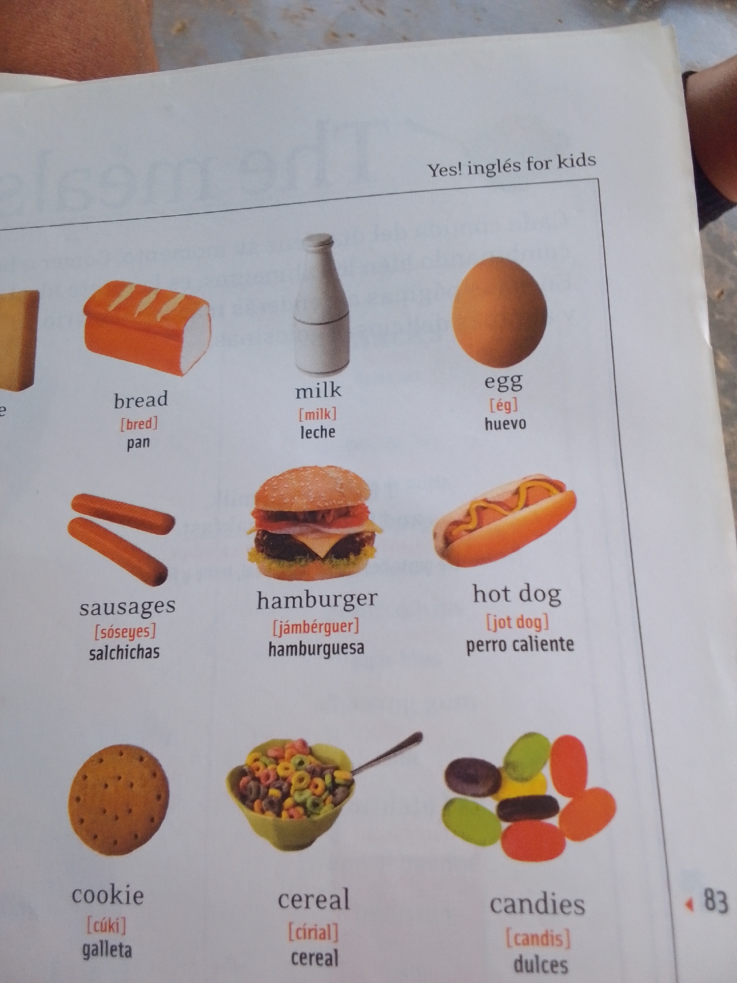 The image is a photograph of an open page from a children's learning book, likely a vocabulary lesson on food items. The page displays colorful illustrations of various foods alongside their English names, phonetic pronunciations, and Spanish translations.
The foods depicted are: bread, milk, an egg, sausages, a hamburger, a hot dog, cereal, and candies. Each item is presented in a clear, simple illustration. The text accompanying each illustration is clearly legible. The page is also visible at the top right, with a title that reads "Yes! inglés for kids". The bottom right corner shows the page number "83".
The overall setting appears to be a bright, indoor environment, suggested by the lighting and the white background of the page. There are no people visible in the image. The activity taking place is a learning exercise, presented through a visual aid. No specific time of day, weather, or location cues beyond the implied educational context are present. The mood is neutral and informative.