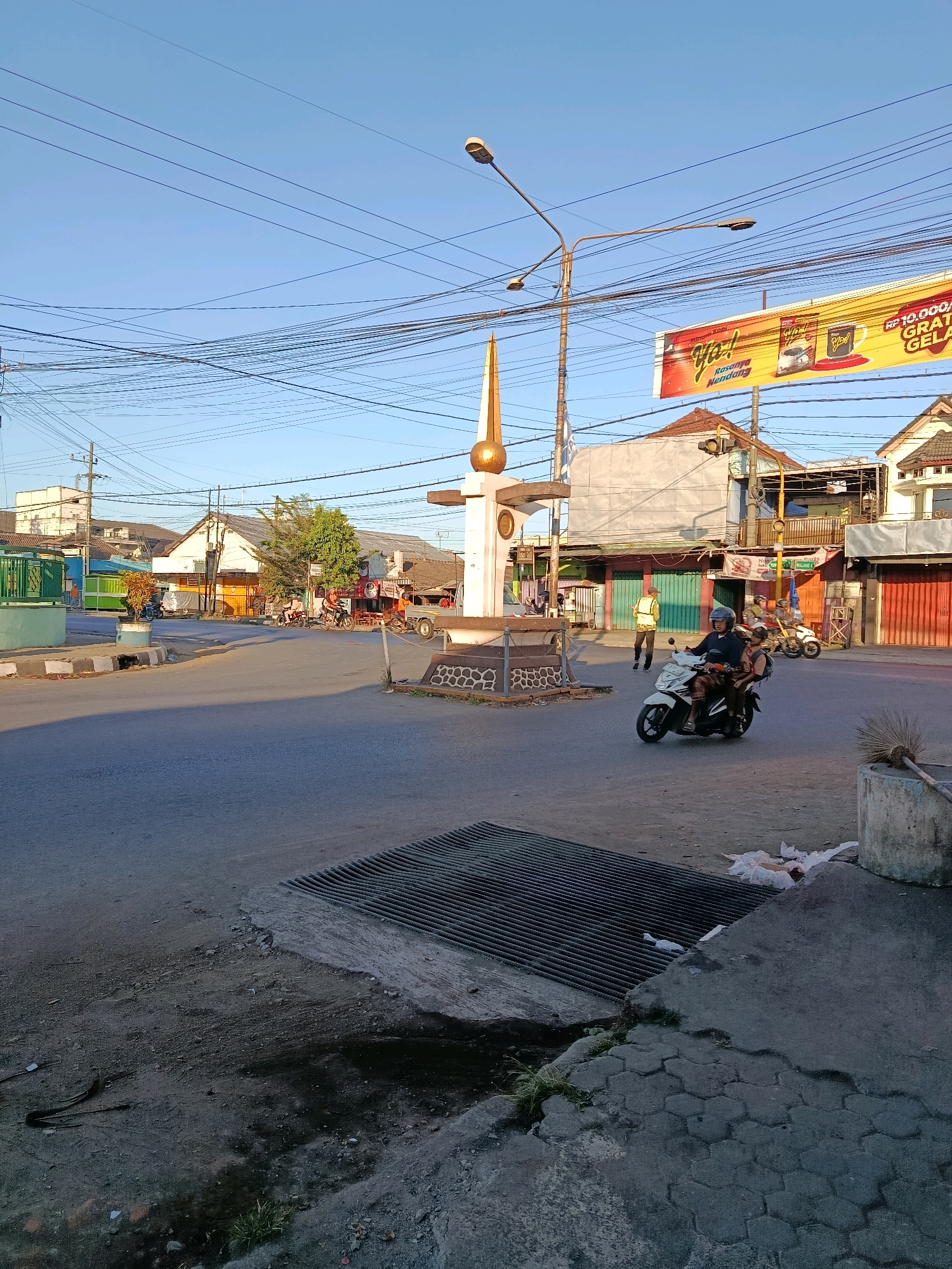The image captures a street scene in Bululawang, Indonesia, bathed in the warm light of what appears to be late afternoon or early morning. The sky is a clear, pale blue. In the foreground, a wide asphalt road curves through the frame. A prominent metal grate is embedded in the pavement on the right side, with some dark, damp earth and a scattering of white debris near it. A rough-textured concrete pillar stands to the right of the grate, with a broom leaning against it.
Dominating the mid-ground is a traffic island with a monument in its center. The monument is a white obelisk topped with a golden sphere. Surrounding the base of the obelisk is a low, decorative stone wall.
Several people and vehicles are present. On the right side of the island, a white motorcycle carries two passengers, a man driving and a woman behind him, both wearing casual clothing. Further back, on the same side of the road, a person in a yellow vest stands near a building. To the left of the monument, several motorcycles are parked, and two people are riding motorcycles heading away from the camera.
The background reveals a street lined with buildings. On the left, there's a green fence and a light blue building. Further down the street on the left, more buildings with light-colored facades are visible. On the right side of the road, there are buildings with tiled roofs, some with colorful awnings and shops. A large yellow billboard advertising "Minum Kopi Ya!" with images of coffee products hangs above the street. Overhead, a complex network of electrical wires crisscrosses the sky, supported by poles. Traffic lights are visible above the street. The overall impression is a busy, ordinary day in a town.