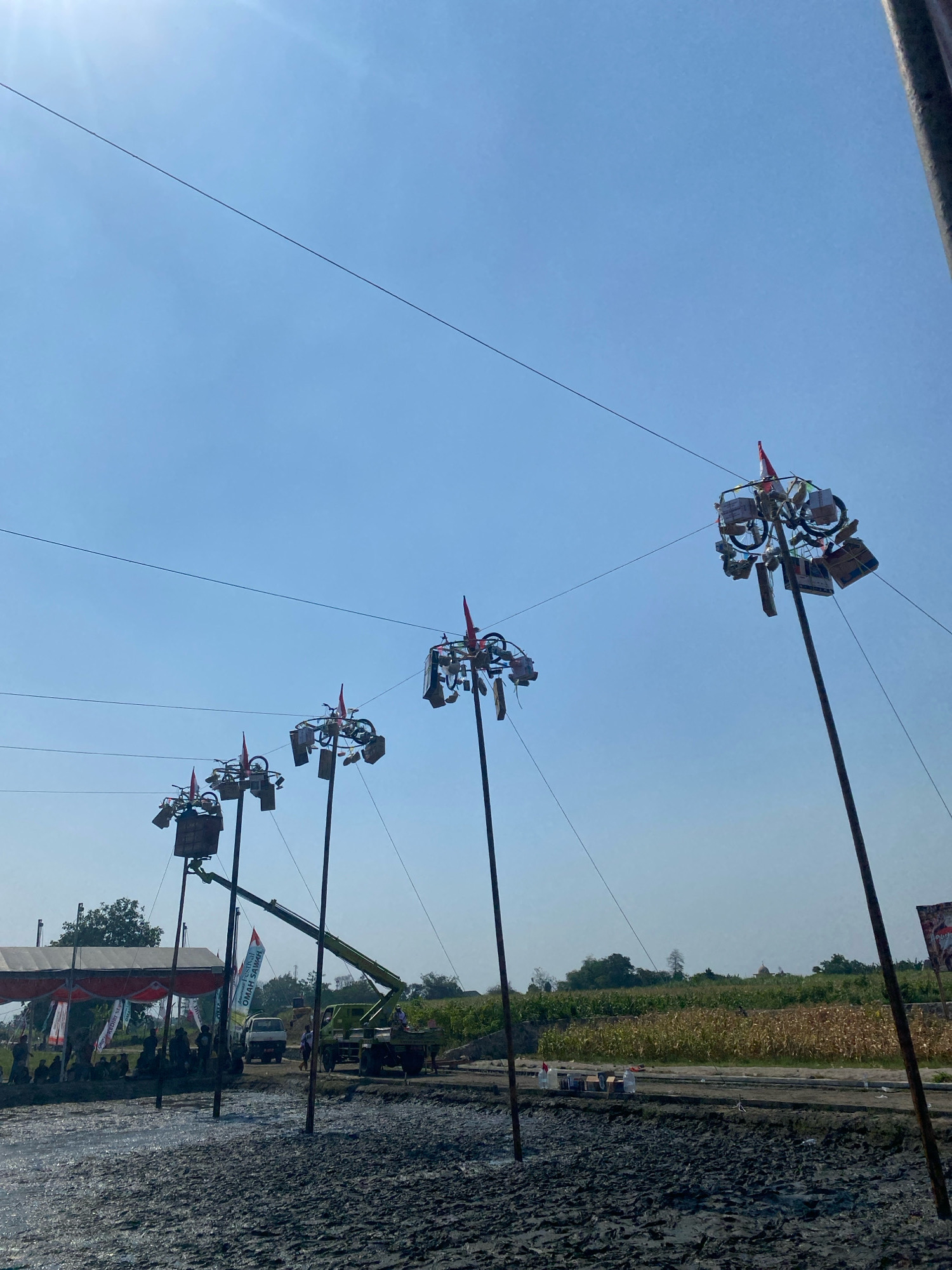 The image depicts an outdoor scene in Kediri City, Indonesia, under a clear blue sky. In the foreground, there is a muddy or wet ground area. Several tall poles are erected, each topped with decorations, including what appears to be bicycles, flags, and boxes. These poles are arranged in a line, stretching towards the background.
In the middle ground, there are tents with red and white awnings, and a gathering of people is visible underneath them. A green crane is positioned behind the tents, with its arm extended upwards. A truck is also present near the crane. In the distance, there is agricultural land with rows of crops, and some trees. The overall atmosphere suggests an outdoor event or festival preparation.