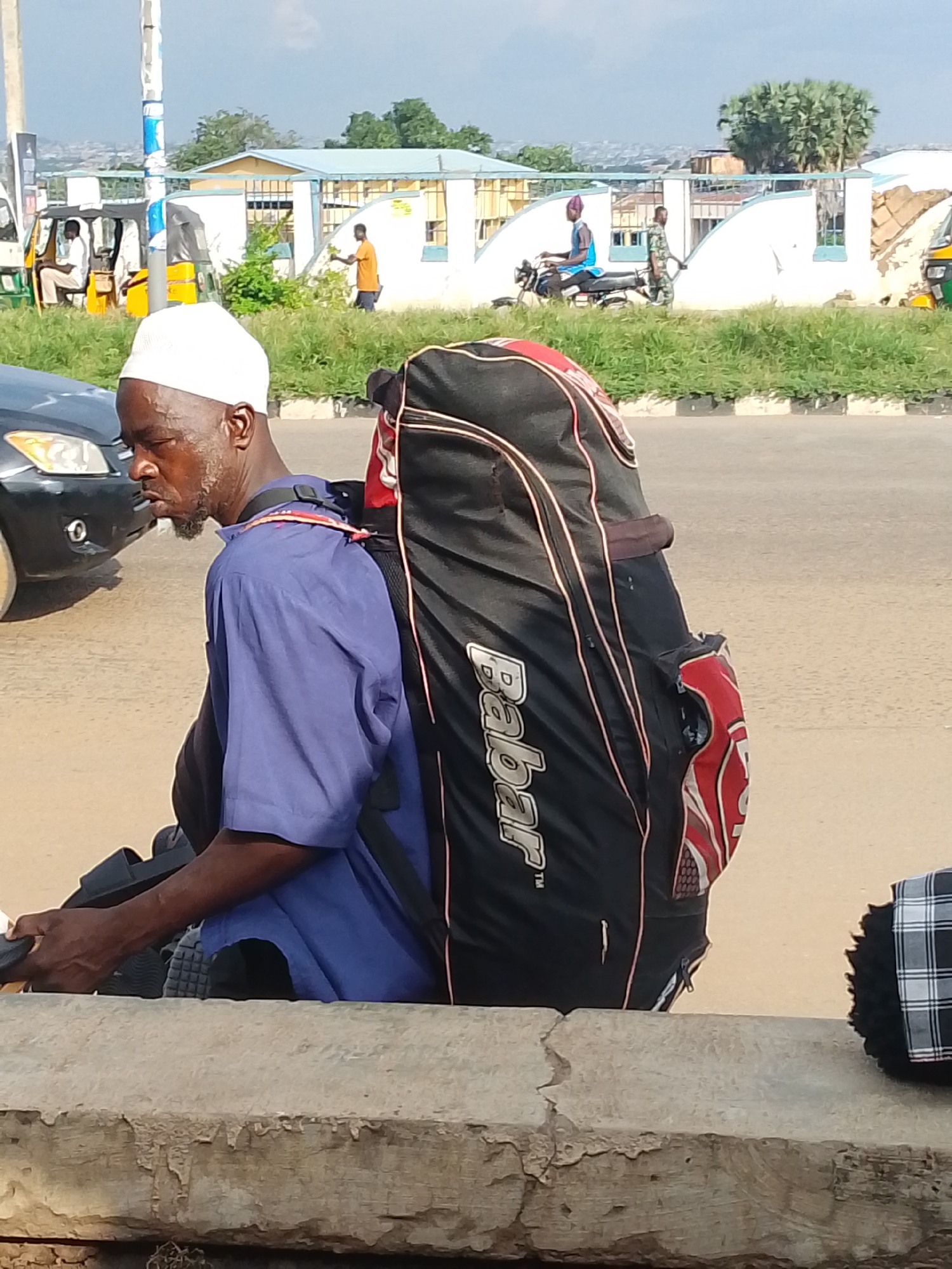 A man wearing a white cap and a blue shirt is riding a motorcycle and carrying a large black bag with red accents on his back. He is on a street in Oja Oba, Oko Erin, Nigeria. The street appears to be paved, and there is a raised concrete barrier in the foreground. In the background, there are buildings, trees, and other vehicles, including a tuk-tuk and other motorcycles. The sky is partly cloudy.
