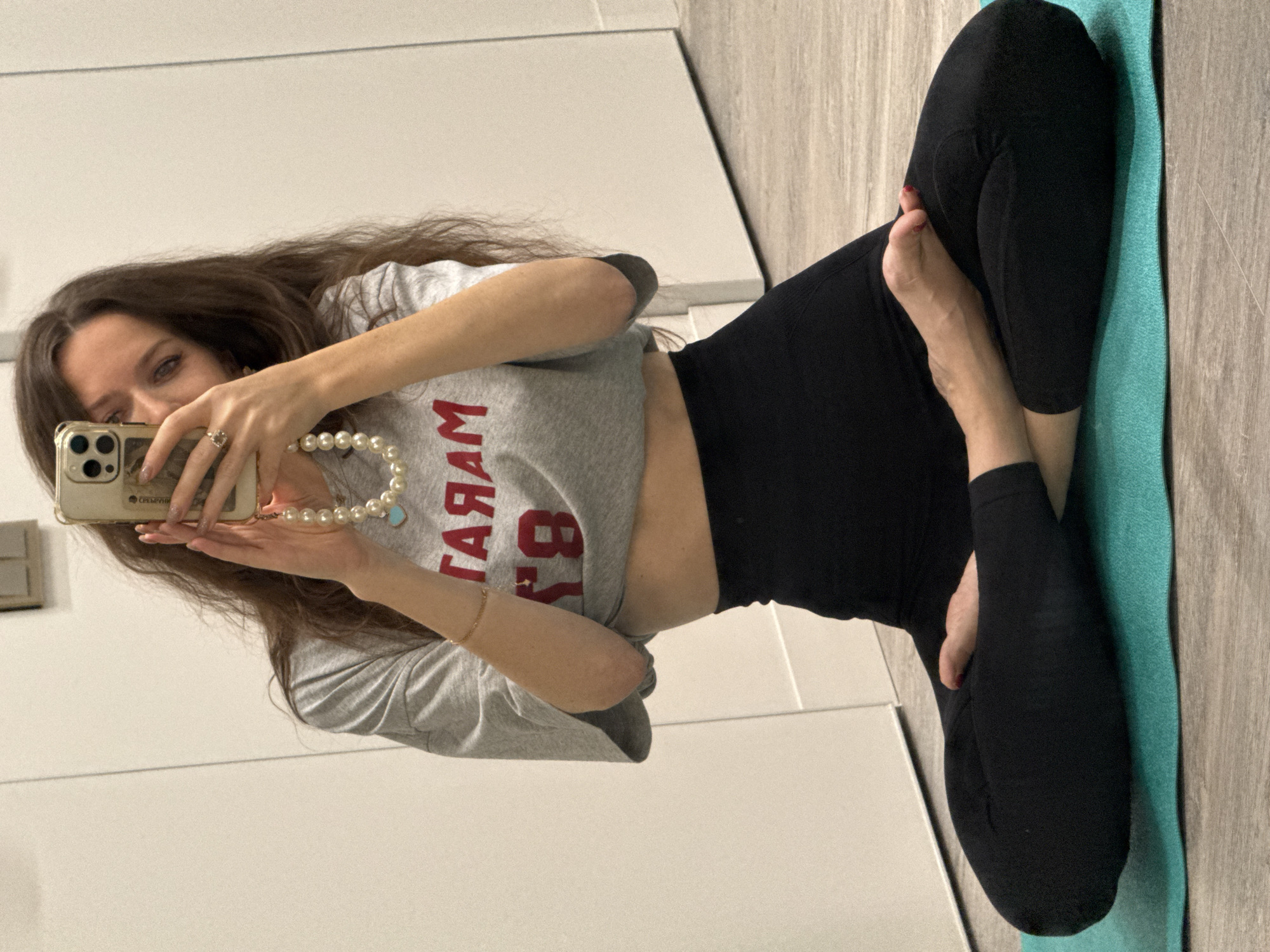 A woman is centered in a photograph. The woman is seated on the floor on a blue-green yoga mat. The woman is wearing black leggings and a grey crop top with red lettering. She is holding a gold cell phone, with an off-white, pearl-like necklace hanging from her right hand. She is wearing a gold ring on her right hand and a thin gold bracelet on her left. She has long, brown hair. The background of the image is primarily a white wall, with brown and grey wood flooring on the right. The location in the city of Unknown, Unknown.