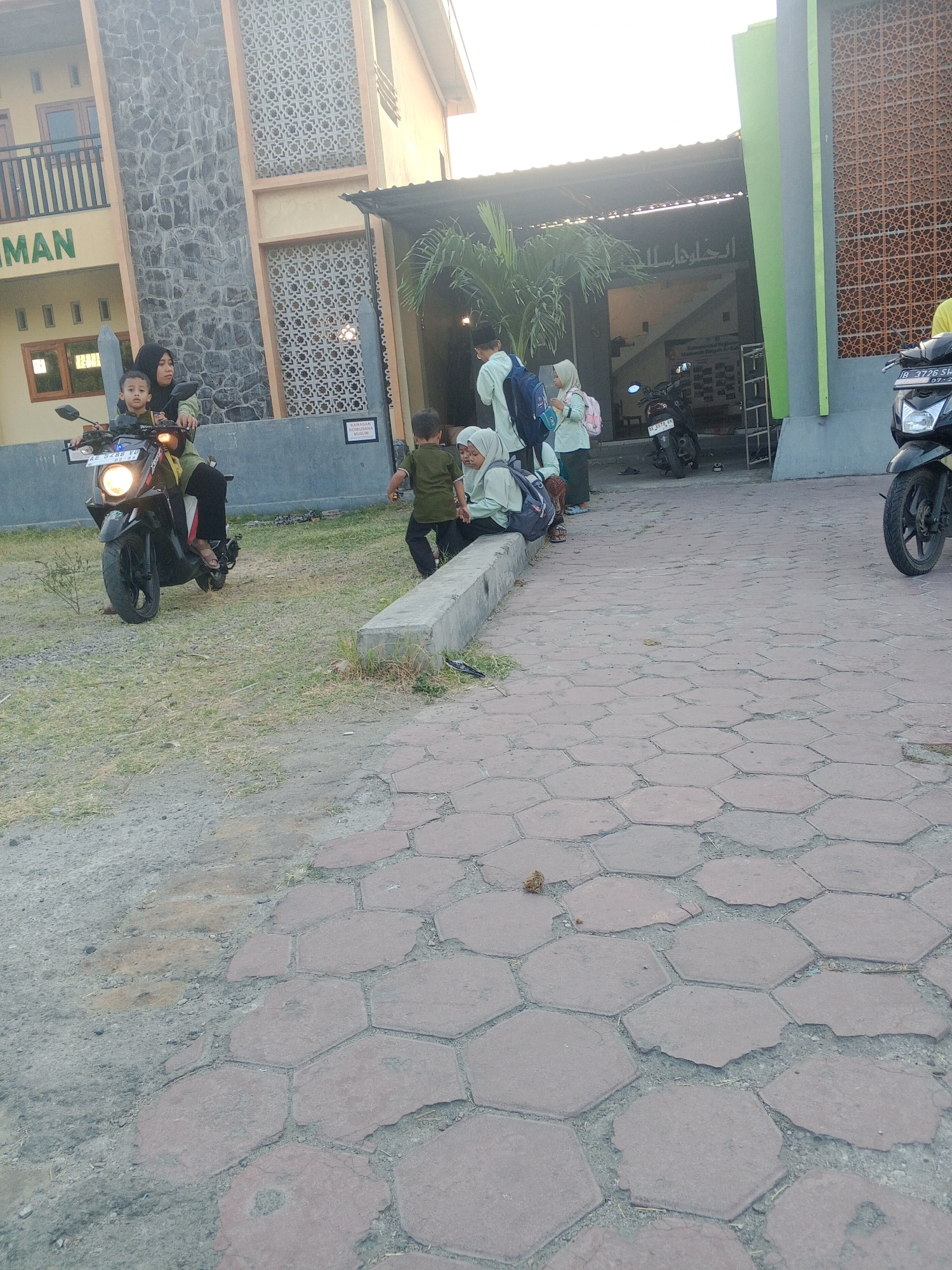 The image shows an outdoor scene in Lembah, Indonesia. In the foreground, a paved area with hexagonal stone tiles is visible, with some dirt and grass growing in the gaps. In the middle ground, a woman wearing a hijab rides a scooter with a young child sitting in front of her. The scooter is black and has a license plate. Behind them, a group of children, some wearing hijabs and school uniforms, are gathered near a building. One child is holding a backpack. The building appears to be a mosque or educational institution, with Arabic script visible above an entrance. A palm tree is also present near the building. In the background, another scooter is parked. The lighting suggests it is daytime, possibly late afternoon due to the warm tones.
