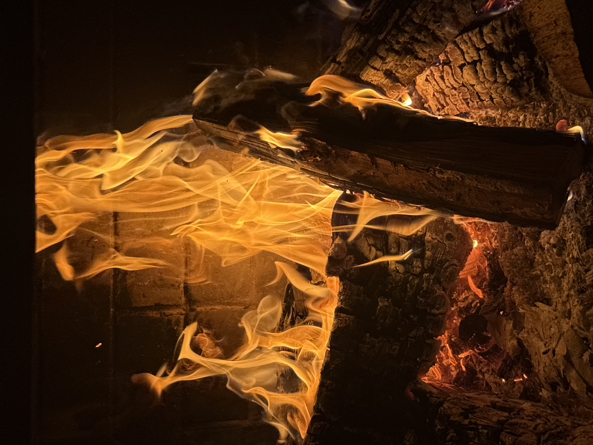 This close-up image captures a vibrant, roaring fire within what appears to be a fireplace or wood-burning stove. The primary subjects are the intense, bright orange and yellow flames and several actively burning, charred wooden logs.
The flames dominate the left and central portions of the frame, swirling and dancing with a luminous quality. They cast a warm, flickering glow across the entire scene. Multiple logs, blackened and deeply textured from combustion, are arranged within the firebox. One prominent log angles downwards from the upper right, its surface showing rough bark and deep cracks, glowing with embers beneath its edge. Another log lies more horizontally, partially consumed by the fire, with flames enveloping its sides. The intense heat has caused the wood to crack and splinter, revealing glowing red-orange embers deep within the structure of the logs.
In the background and to the left, faint brickwork is visible, indicating the interior of a fireplace. The overall setting is dark, with the fire serving as the sole light source, creating a high contrast scene that emphasizes the fire's warmth and energy. Wisps of smoke are subtly present, mingling with the flames.
The activity taking place is the active burning of wood, creating light and heat. The scene evokes a sense of warmth, coziness, and tranquility, typically associated with a fire lit during the night or a cold period. No people or visible text are present. The location, as per context, is Заимка (снт), Russia, suggesting a rural or suburban home setting.