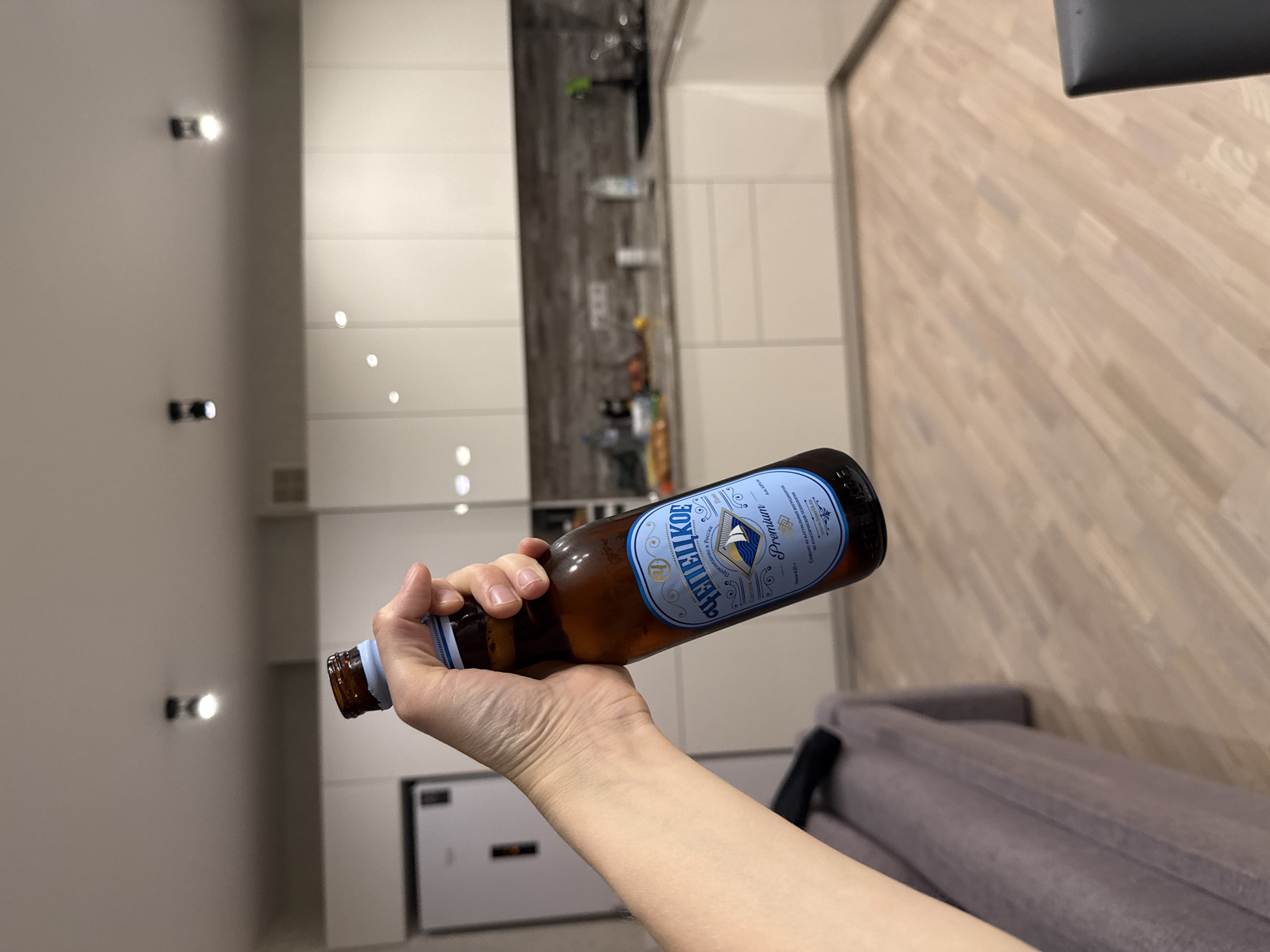 A person is holding a brown glass bottle of "Chepetskoe Premium" beer in their hand, with the label facing the viewer. The bottle has a blue and white label with gold accents and Cyrillic text. The hand appears to be fair-skinned.
The background includes a modern interior with white cabinets, recessed lighting, and a light-colored wood floor. A gray sofa is partially visible in the lower right corner. Based on the label text "Сделано в России" (Made in Russia), the beer is Russian. The beer is produced in the city of Izhevsk in Russia.