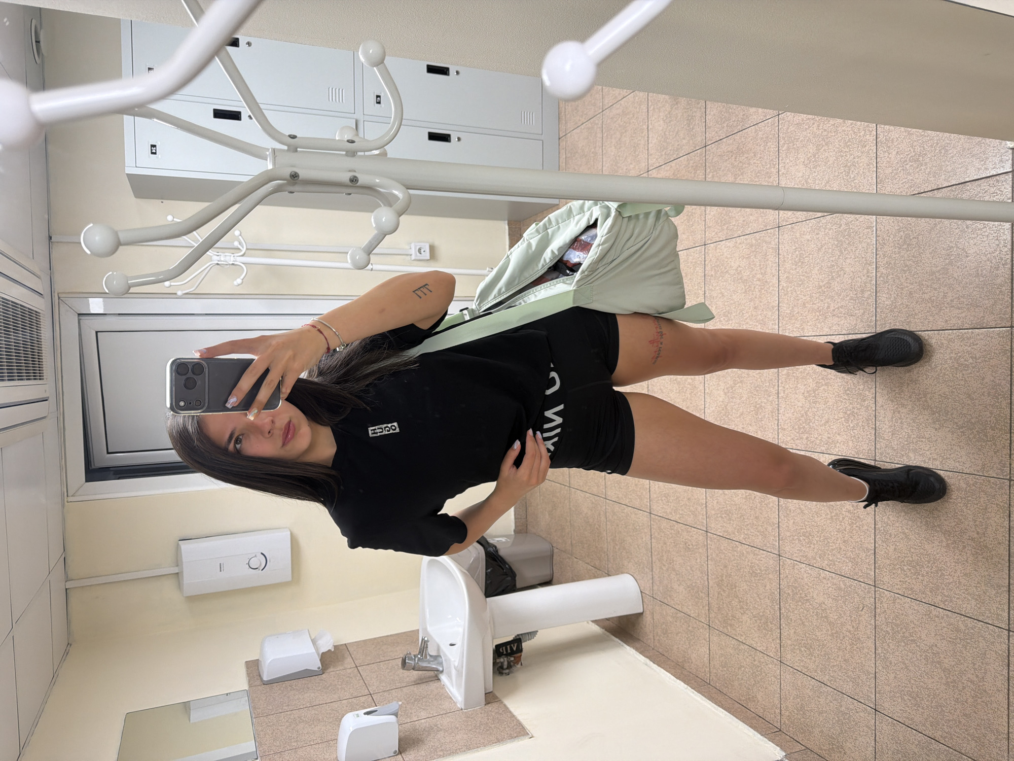 A young woman takes a selfie in a restroom in Gabrovo, Bulgaria. She stands in front of a mirror with a black shirt, black shorts, and black sneakers. Behind her is a sink, soap dispenser, paper towel dispenser, and a coat rack with lockers in the background. The floor is tiled.