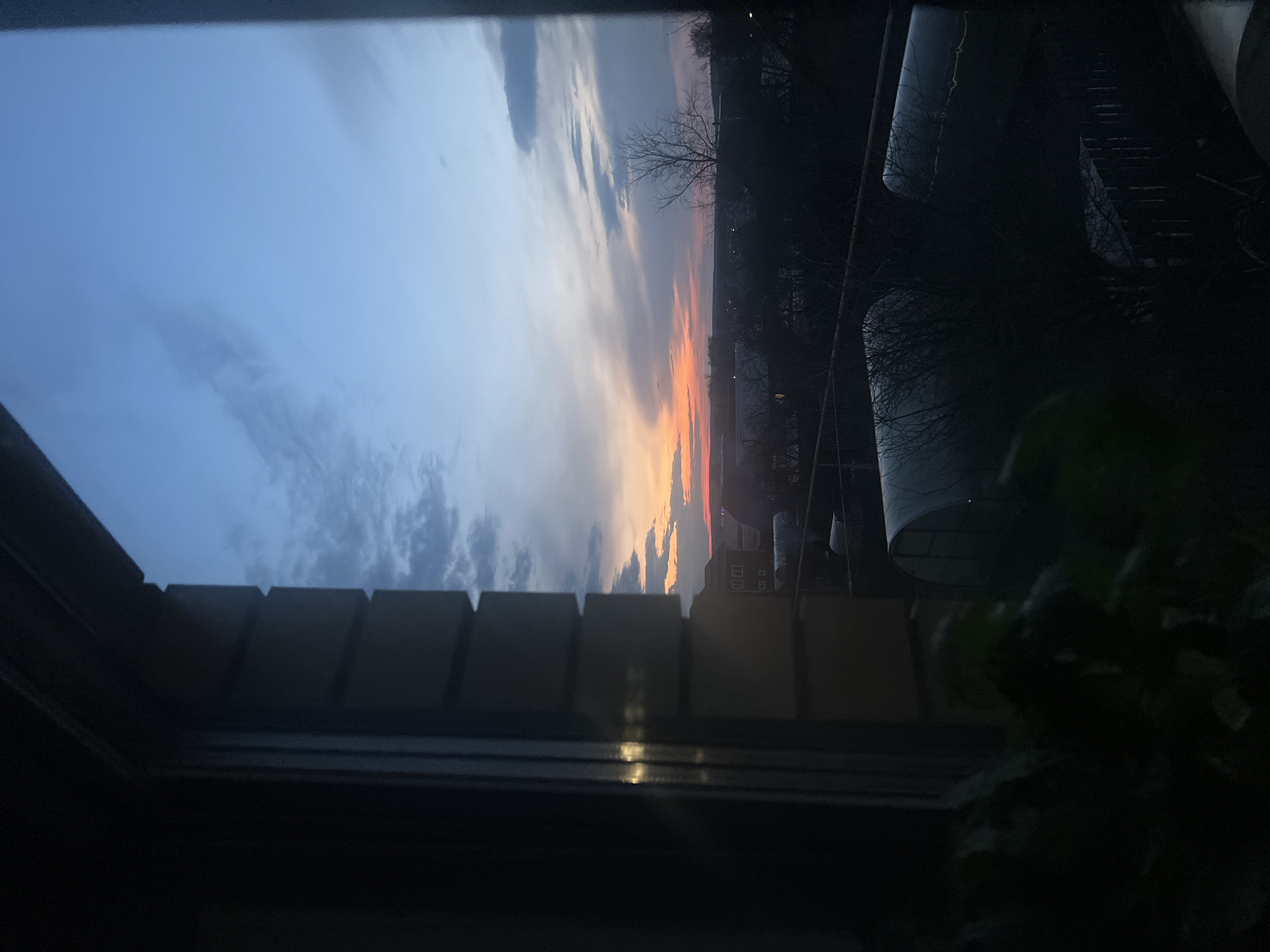 The image shows a view of the sky at sunset from a window. The sky is a gradient of colors, transitioning from blue to orange. Clouds are visible in the sky. Outside the window, there are trees and buildings. The window frame is visible in the foreground, partially obscuring the view. The image appears to be taken from an indoor setting. There are no people visible in the image. The location in the city of Votkinsk, Russia is not specified, therefore no conclusions can be made to confirm if the image was taken there.
