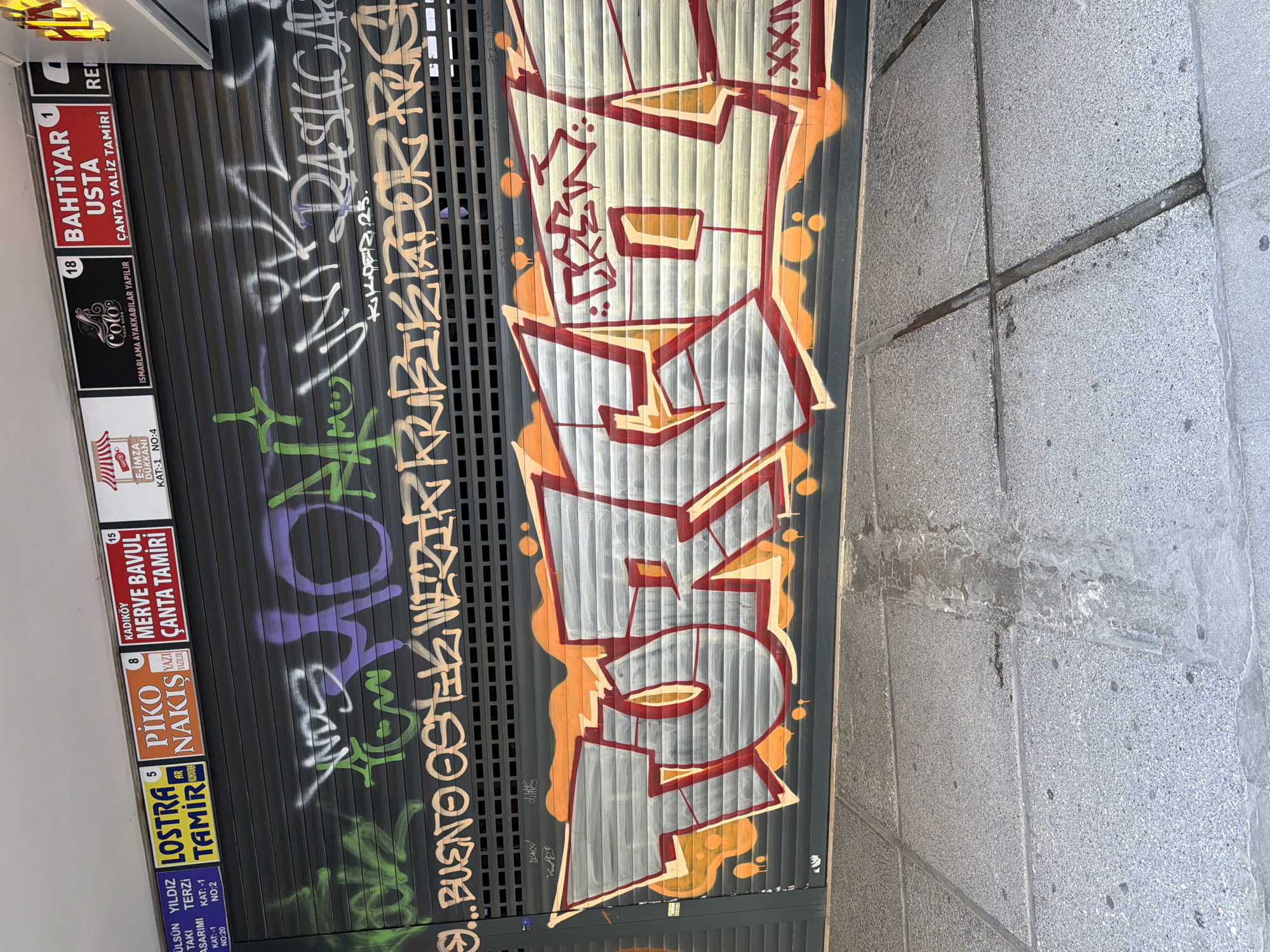The image shows a close-up view of a metal security shutter in the Kadıköy district of Istanbul, Turkey, covered in graffiti. The shutter is gray and has horizontal grooves. The graffiti includes large, colorful bubble letters that appear to spell out "TORKIZ" in red and white with orange and yellow accents. Above this, there is smaller, lighter-colored graffiti that is difficult to read, including some green spray paint. The top of the image features several small signs, likely for businesses. One sign is red and says "BAHTİYAR USTA ÇANTA VALİZ TAMİRİ" (Bahtiyar Master Bag Suitcase Repair). Another sign is orange and says "PİKO NAKİŞ YAZILIR" (Pico Embroidery Written). Further to the left, a blue sign reads "YILDIZ TERZİ TAKI TAMİRİ" (Yildiz Tailor Jewelry Repair). The sidewalk in front of the shutter is made of gray concrete slabs with visible lines and some dark stains.
