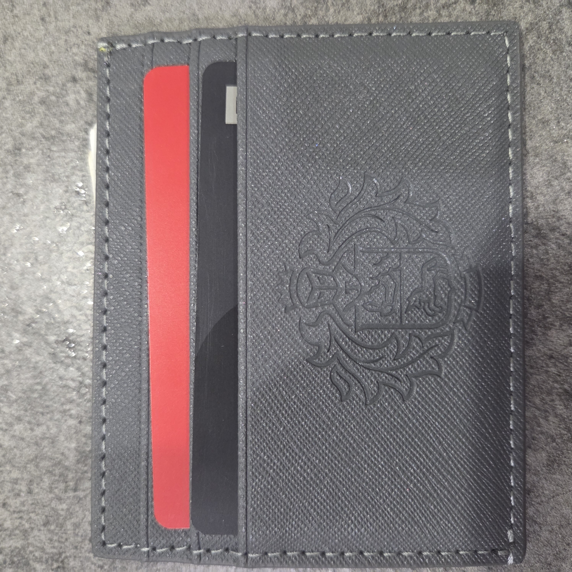 The main image displays a dark grey card wallet or holder, positioned flat on a textured, grey background. The wallet is made of a material with a distinct cross-hatch or Saffiano-style texture. White stitching runs along the edges of the wallet, creating a visible contrast.
Two cards are partially visible in slots on the left side of the wallet: a bright red card is in the leftmost slot, and a black card with a narrow, vertical white rectangular element is in the adjacent slot to its right.
On the right side of the wallet, an intricate, stylized design or logo is embossed into the material. It appears to be a crest or emblem with swirling, decorative elements.
The background surface is grey and textured, possibly a stone or fabric, with several small, irregular, lighter-colored, reflective specks scattered across it, particularly on the left side. No people are present, and no specific activity or event is taking place. There is no discernible text on the wallet or cards that can be read. The time of day, weather, and specific location cues are not apparent from the image.