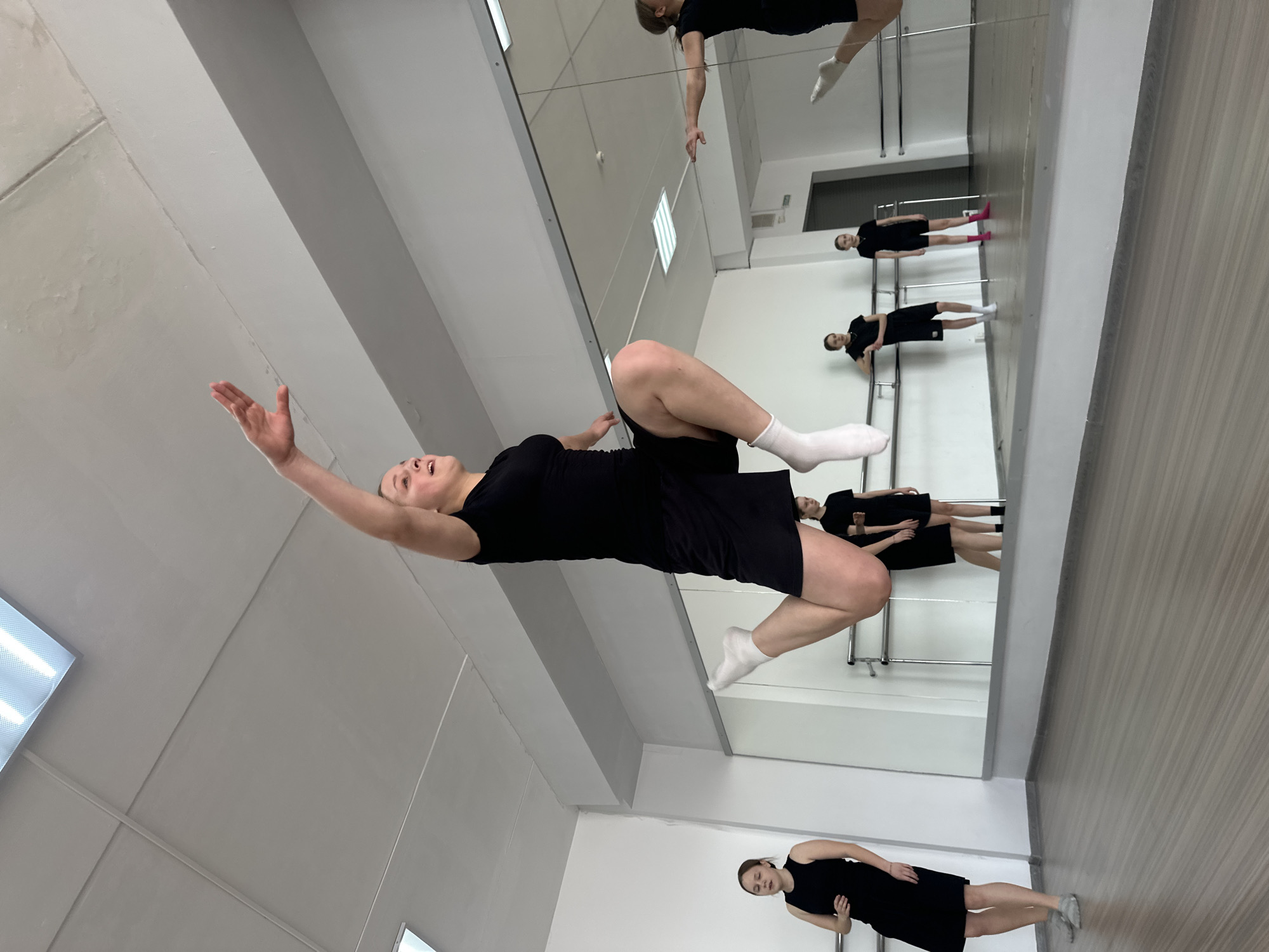 The image shows a dance studio in Votkinsk, Russia. A woman in a black dress and white socks is captured mid-air, performing a dance move. She is facing upwards with her right arm raised. The studio is mirrored, creating multiple reflections of the dancer. Other women in similar attire are seen either standing or practicing near the mirror. The studio has a light-colored floor and ceiling, with fluorescent lighting. A ballet barre runs along the wall near the mirror.