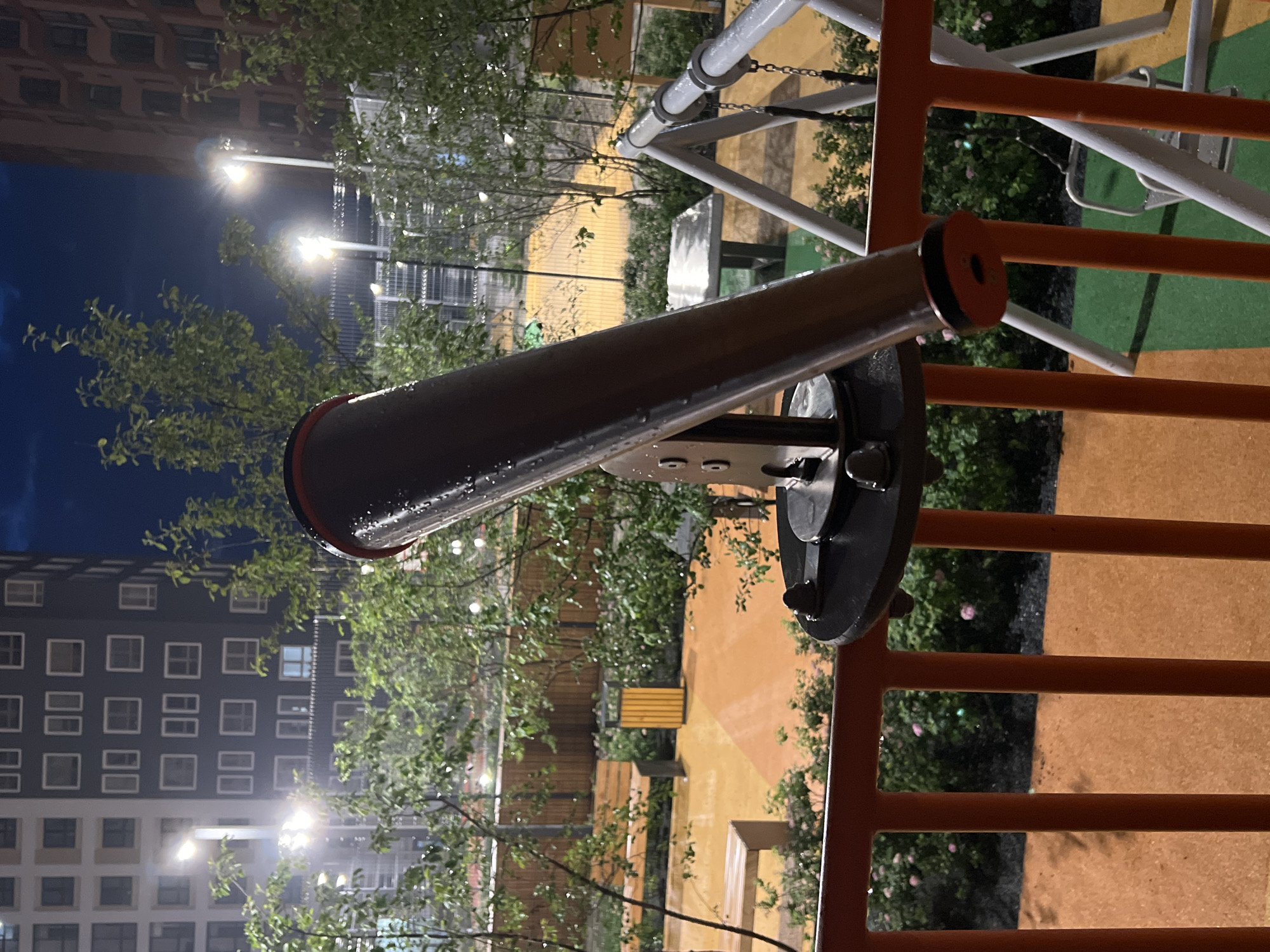 A telescope is mounted on a circular base with a metal arm, positioned in the foreground on an orange railing. Behind the railing is a playground with an orange and green surface, a swing set, benches, a trash bin, and trees. In the background, two apartment buildings are visible, illuminated by streetlights. The sky is dark blue, suggesting it is nighttime. There is no information to determine the city of Yekaterinburg, Russia.