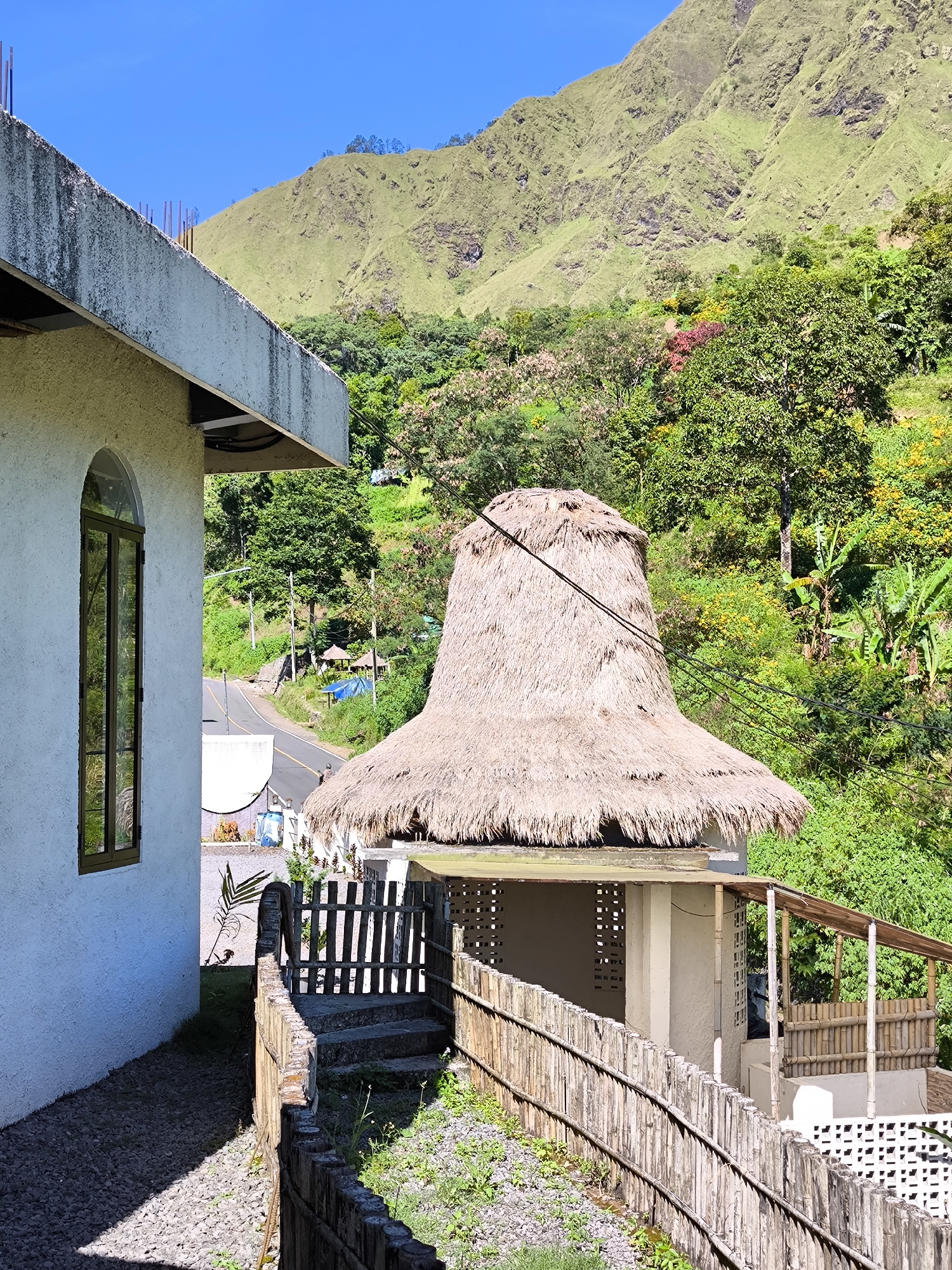 The photograph captures a rural scene in Sembalun Bumbung, Indonesia. The foreground is dominated by a white stucco building with a tall, arched window. Adjacent to the building is a wooden fence that runs along a stone path leading to a small structure with a thatched roof resembling a traditional Indonesian hat. The thatched roof structure has open sides with bamboo detailing and a raised floor. In the background, a lush green hillside rises steeply, covered in trees and vegetation. A road is visible winding through the trees, and a few small structures can be seen in the distance. The sky is bright blue, suggesting a clear day. Electrical wires are visible running across the scene, cutting through the natural landscape.
