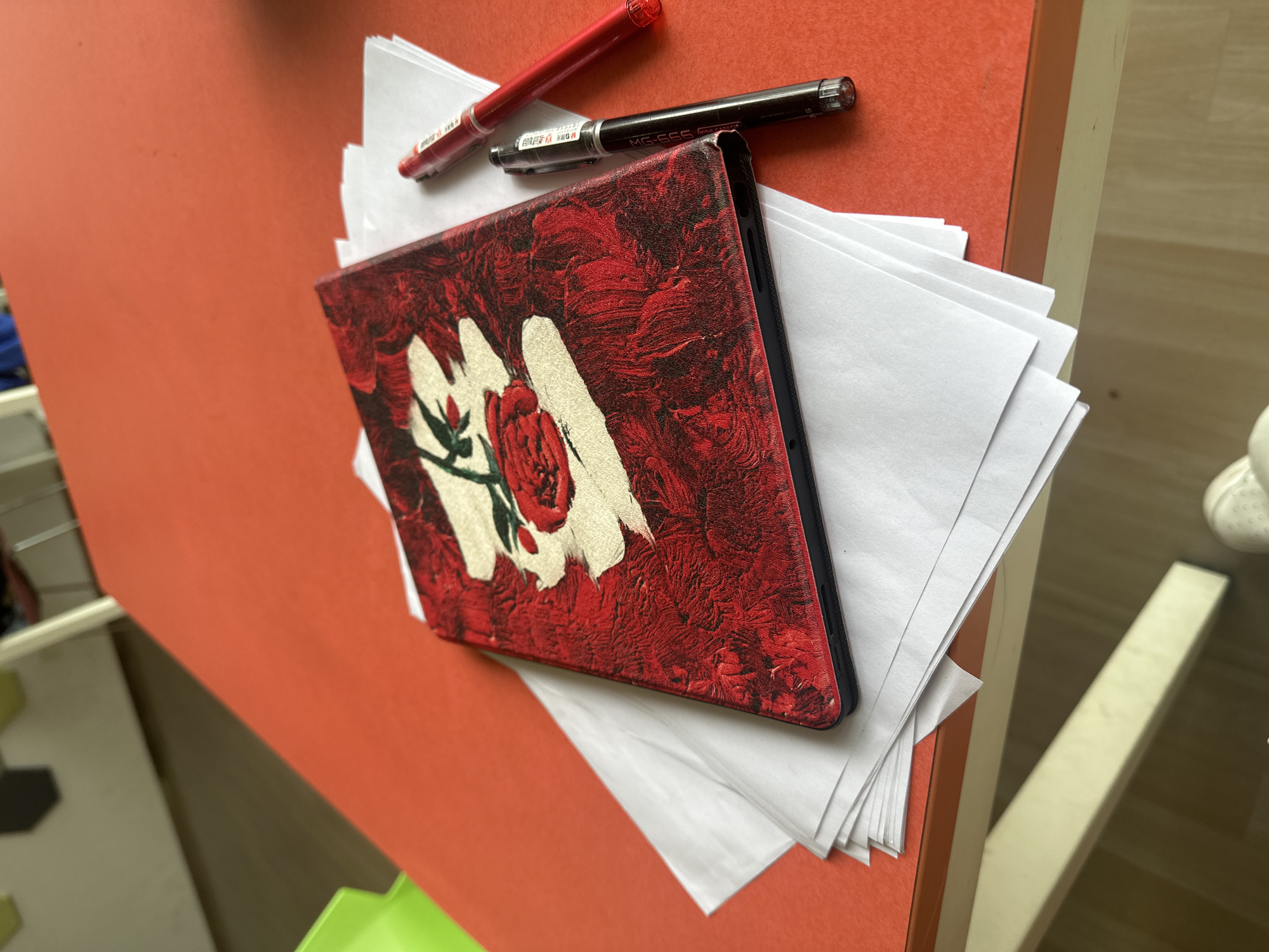 The image shows an orange table or desk surface with a stack of white paper sheets. On top of the paper are two pens: one red and one black. A tablet or book with a red textured cover with a white and red rose design is positioned centrally. The items are located in Chongqing High-tech Zone, China. The setting appears to be indoors.
