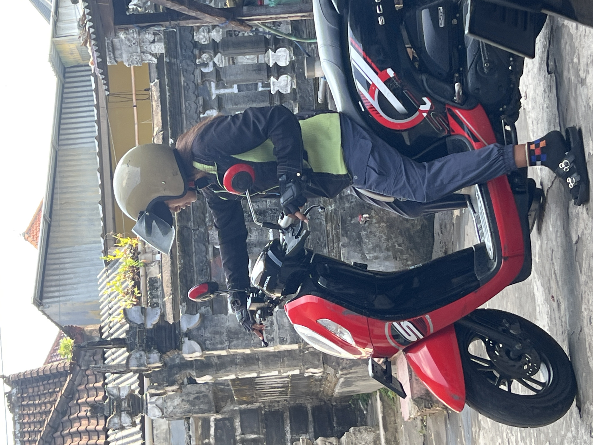 A person wearing a helmet, jacket, and pants is seated on a red and black scooter. The person is holding the handlebars. The scooter is parked on a paved surface in front of a building with a tiled roof and a stone wall. The environment suggests a street scene in Denpasar, Indonesia. The person's socks have a checkered pattern of orange, blue, and white. The scooter has a Honda logo on the front.