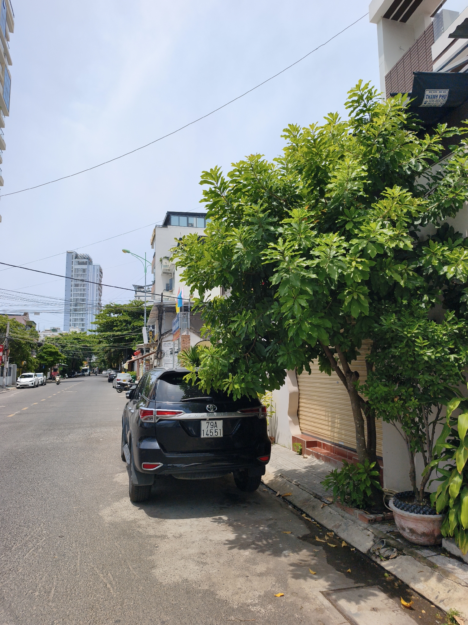 A black SUV is parked on the side of a street in Nha Trang, Vietnam. The vehicle has a license plate that reads "79A 145.51". In the background, there are several buildings, including a tall, modern skyscraper. Trees line the street, and the sky is clear and blue. A few other cars are visible on the road, as well as some power lines. The scene appears to be a typical day in a bustling Vietnamese city.