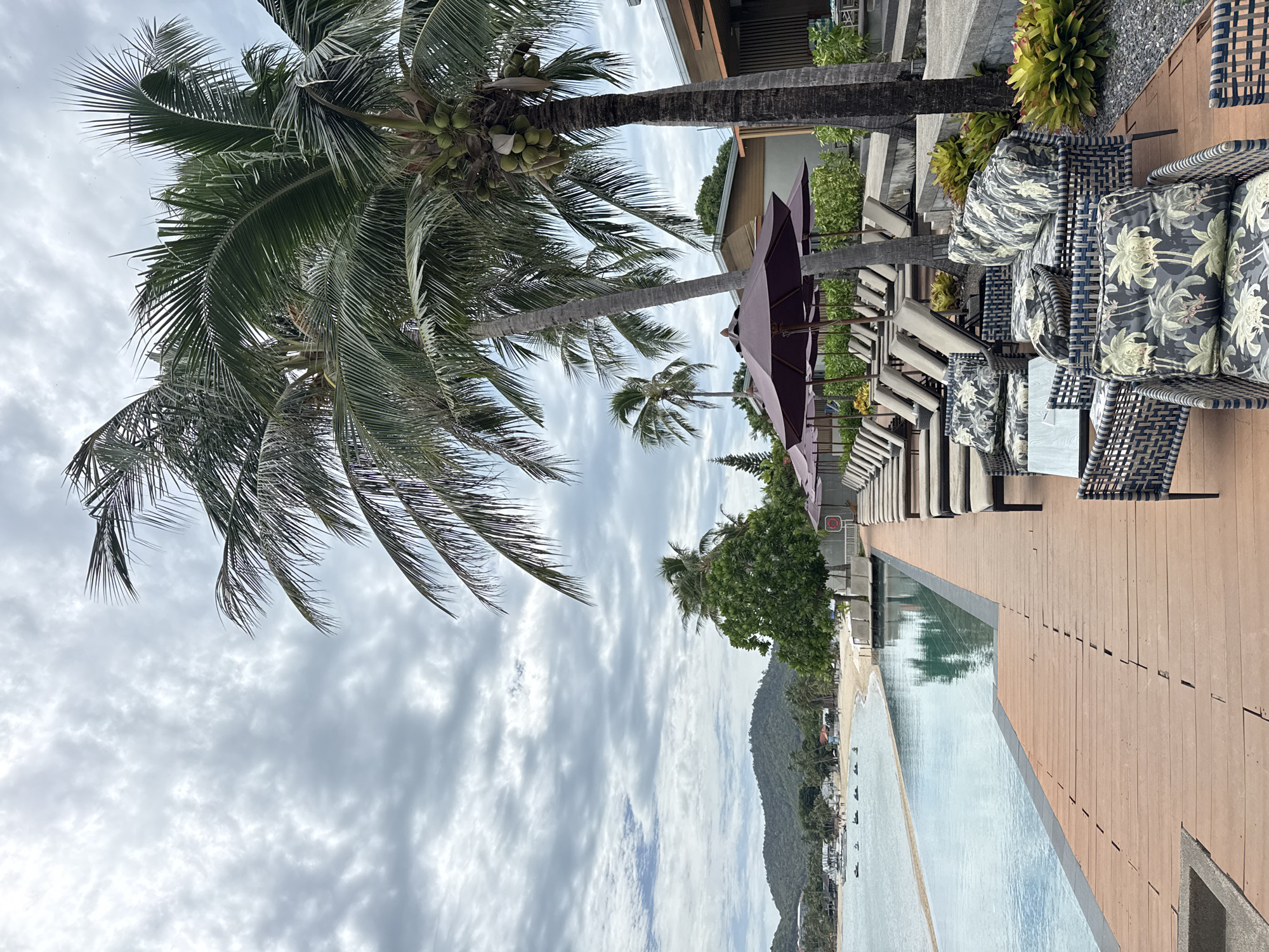 The image depicts a tropical resort scene in the Ko Samui District of Thailand. In the foreground, a wooden deck leads to an infinity pool, which stretches towards a sandy beach and the ocean. Palm trees with coconuts are visible, as are several lounge chairs with dark umbrellas. In the middle ground, the beach is lined with more palm trees and some resort buildings. The background features rolling green hills under a cloudy sky. Several jet skis are visible on the water. The overall impression is one of a serene, luxurious vacation spot.