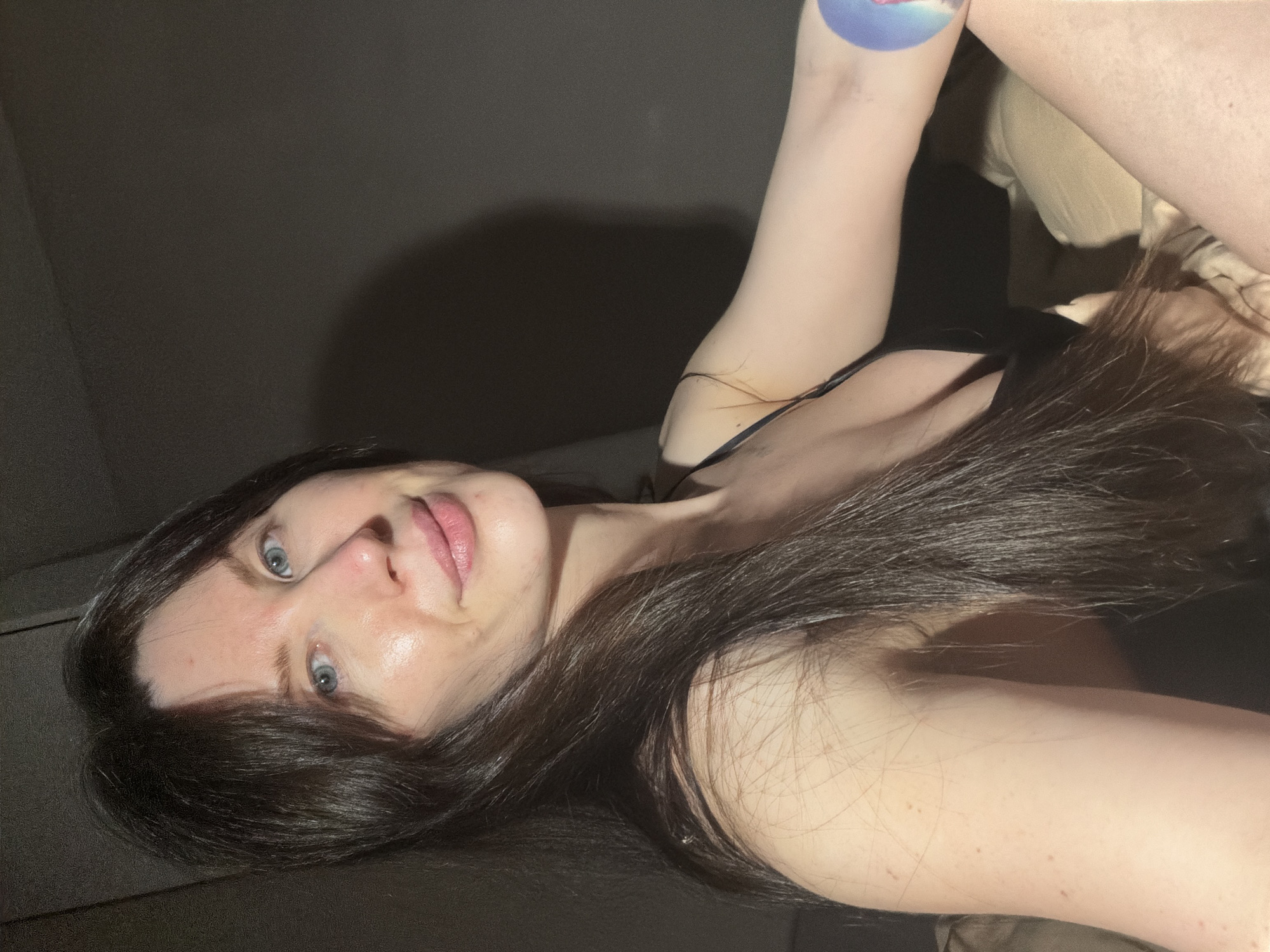 The image shows a woman with long, dark hair and fair skin. She is wearing a black top with thin straps. Her eyes are light-colored. Part of her arm is visible, with a round sticker on it. The background is dark, suggesting a dimly lit room. The image doesn't provide any information about the location in the city of Perm, Russia. The image appears to be a selfie.