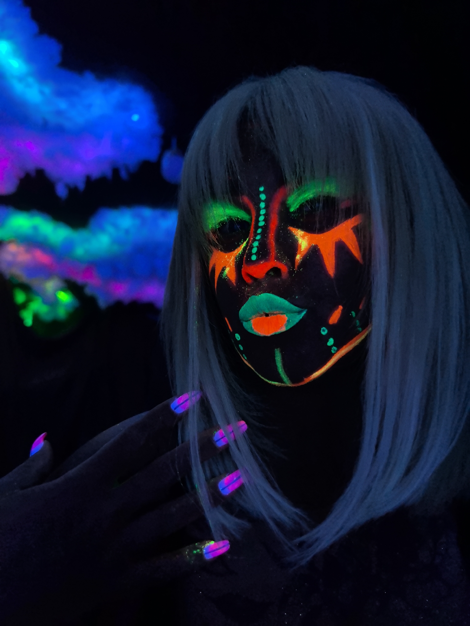 A striking, close-up portrait captures a person illuminated by ultraviolet light, showcasing elaborate glow-in-the-dark face paint and accessories. The subject wears a light, possibly silver-blonde, shoulder-length wig with bangs. Their face is adorned with intricate patterns of neon orange, green, and red paint; prominent features include orange triangular shapes above the eyes, green lines around the eye sockets, a red accent on the nose, and lips painted with a vibrant red center and a green outline. Additional green lines and dots decorate the cheeks and chin.
One hand is visible in the lower left, with fingers gently splayed. The fingernails are long and painted with a dual-tone neon pink and blue gradient, glowing intensely under the UV light. The background is completely dark except for large, abstract, cloud-like formations that fluoresce in shades of blue, purple, and green, suggesting an indoor setting specifically designed for blacklight effects. The overall atmosphere is artistic and otherworldly. There is no discernible text. The scene is set in a dark environment, likely at night or indoors with controlled lighting.