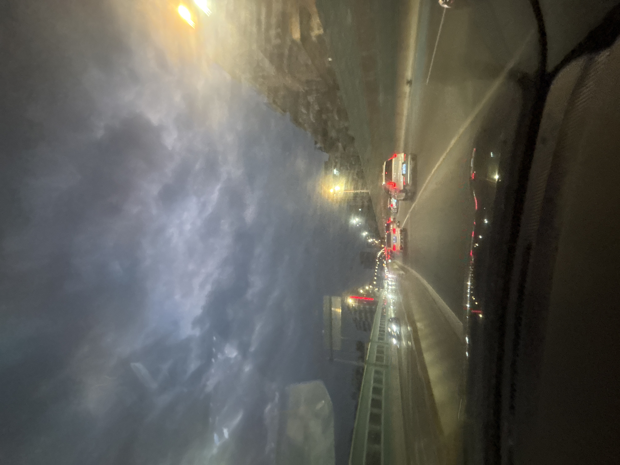 The image captures a view from inside a vehicle driving on a highway in Baiyun District, China, during what appears to be dusk or evening. The sky is overcast with dramatic, dark clouds, hinting at an impending storm or recent rain. Streetlights and the headlights/taillights of other vehicles illuminate the wet roadway, reflecting on the surface. Several cars are visible on the highway, including a white SUV in the foreground. The scenery includes a raised highway barrier to the left and a grassy, sloped embankment on the right, partially obscured by trees and illuminated by artificial lights. The overall atmosphere is moody and dynamic due to the weather and lighting conditions.