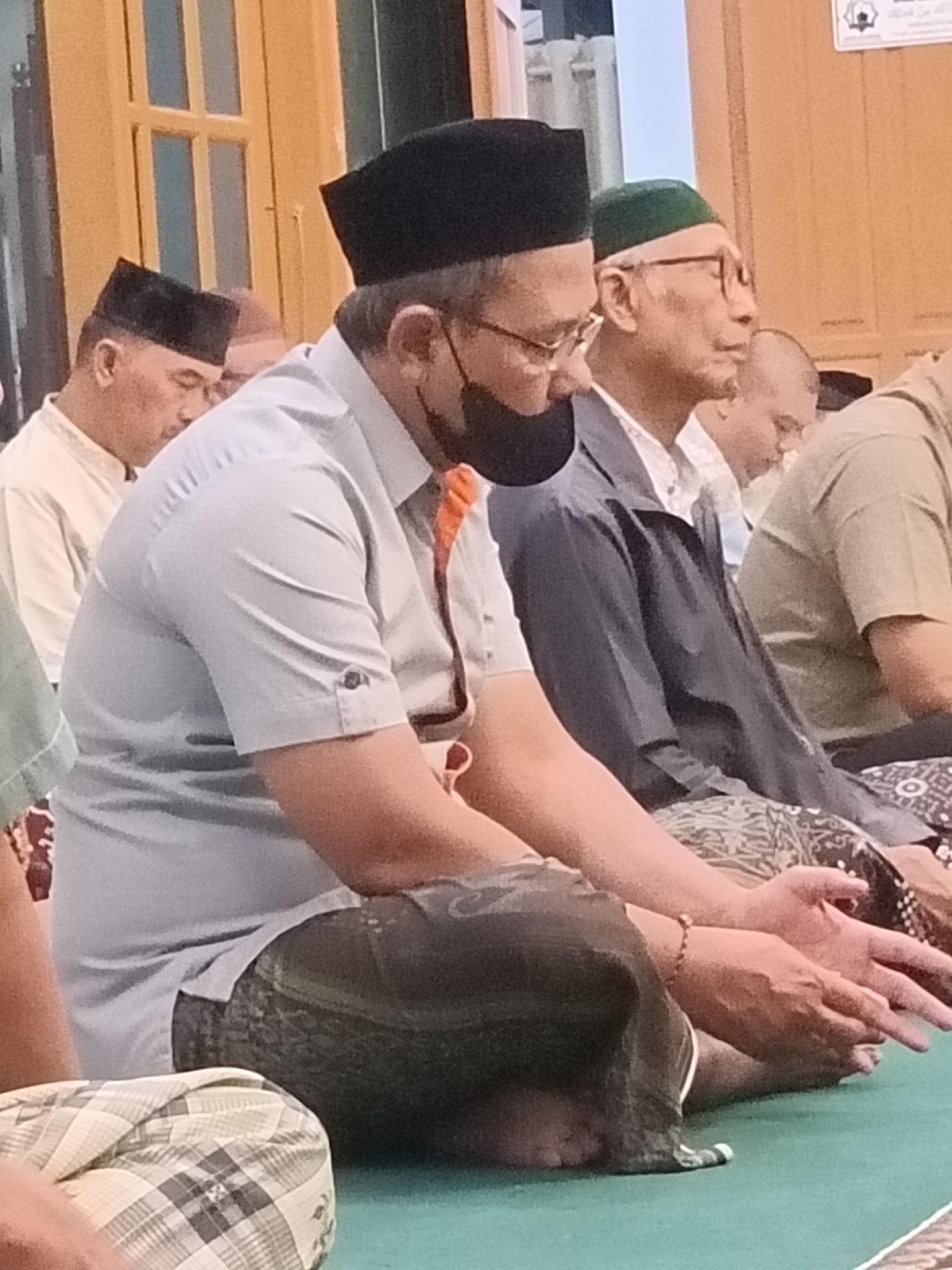 The image depicts a group of men sitting on a green carpeted floor, likely inside a building. The primary subject in the foreground is a man wearing a light blue collared shirt, a black skullcap, and a black face mask. He is seated with his legs crossed and his hands resting on his lap. To his left, another man in a white shirt and black skullcap is partially visible, also seated. Behind the central figure, a man wearing glasses and a green skullcap is visible, looking to his right. Further in the background, other individuals are seated, some wearing skullcaps. The environment appears to be indoors, with wooden paneling on the walls and a window with wooden frames visible on the left. The overall atmosphere suggests a gathering, possibly a religious service or meeting. While the specific location within Kediri City, Indonesia, cannot be determined from the image alone, the attire of the individuals and the presence of skullcaps suggest a cultural or religious context common in Indonesia.
