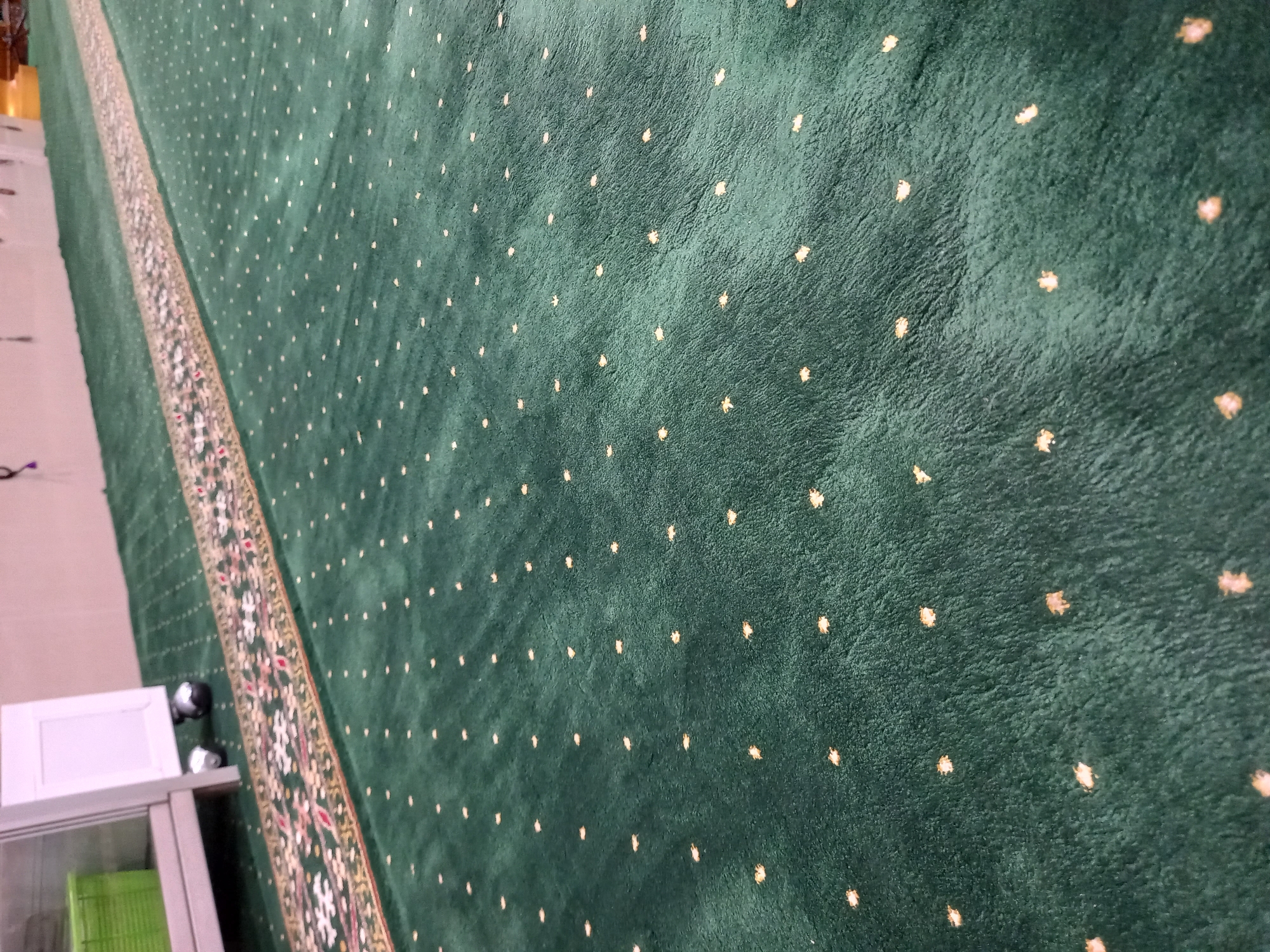 The image shows a close-up, low-angle view of a dark green carpet with a repeating pattern of small, light-colored dots. A patterned runner rug, featuring intricate geometric and floral designs in gold, red, and white, runs diagonally across the frame from the upper left towards the bottom right. In the far upper left corner, a portion of a white cabinet or shelving unit is visible, with a bright green plastic bin inside. The carpeted floor appears to be in an indoor setting, likely a place of worship or a similar public space, given the presence of a prayer rug runner. The lighting suggests an interior environment. The image does not contain enough information to determine its specific location in Samarinda, Indonesia.