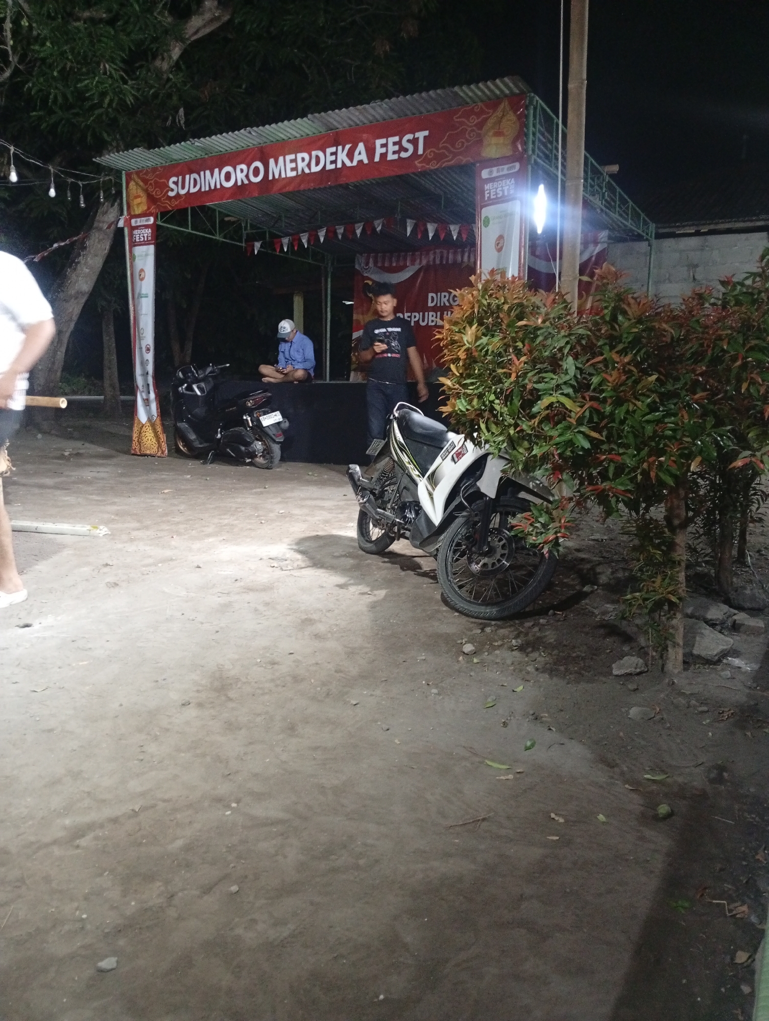The image depicts an outdoor scene at night, likely a festival or event, in Sewon, Indonesia. A red banner with white text reads "SUDIMORO MERDEKA FEST". Underneath the banner, a stage-like structure is visible with decorative triangular flags. Two men are present. One man, wearing a blue shirt and a white cap, is seated on the stage, looking at his phone. Another man, wearing a black t-shirt and jeans, stands on the right, also looking at his phone. Two motorcycles are parked in front of the stage. One is a black scooter-style motorcycle, and the other is a white and black street bike. A bushy plant with red and green leaves is situated to the right of the street bike. The ground is a dirt or gravel surface. String lights are visible hanging above the stage area. The overall impression is of a casual, nighttime gathering.