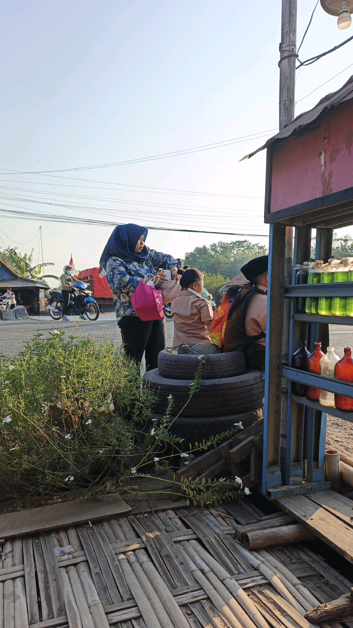 This image depicts a roadside scene in Krimun, Indonesia, with a focus on a small roadside fuel stand. In the foreground, a woman wearing a blue hijab and a patterned shirt stands, holding a pink bag and a mobile phone. She appears to be interacting with two children, who are seated on a stack of tires. Both children are wearing tan school uniforms and backpacks. One child is looking away, while the other faces the woman.
The fuel stand itself is constructed with blue metal framing and has shelves displaying various bottles of colorful liquids, likely gasoline. Red and orange glass bottles are also visible. Behind the fuel stand, a wooden structure, possibly part of the stand's foundation or an adjacent building, is made of bamboo.
To the left of the frame, a motorcycle is seen on the road, with a rider wearing a helmet and a mask. Another person can be seen in the background near a small building. Lush green vegetation with small white flowers grows in front of the tire stack. Power lines are visible crisscrossing the sky, indicating an urban or semi-urban environment. The overall lighting suggests it is daytime, with the sun casting shadows.