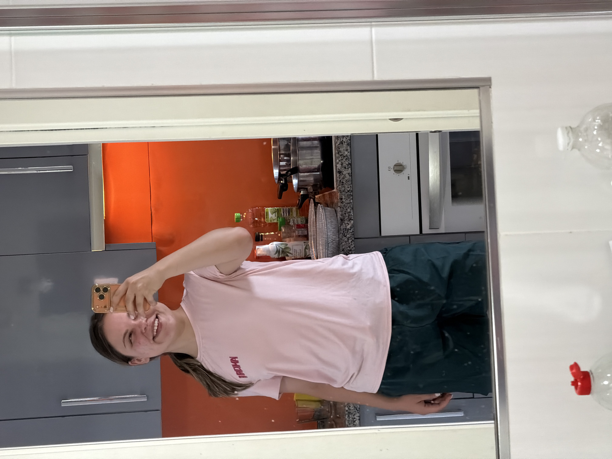 A young woman is taking a mirror selfie in a kitchen. The woman has brown hair and is wearing a pink t-shirt and dark green shorts. She is smiling and holding her phone up in her right hand.
The kitchen has grey and orange cabinets, a stainless steel oven, and tiled walls. There are bottles of oil and other cooking items visible on the counter.
The location is most likely Arroyo de la Miel-Benalmádena Costa, Spain, as specified in the request.